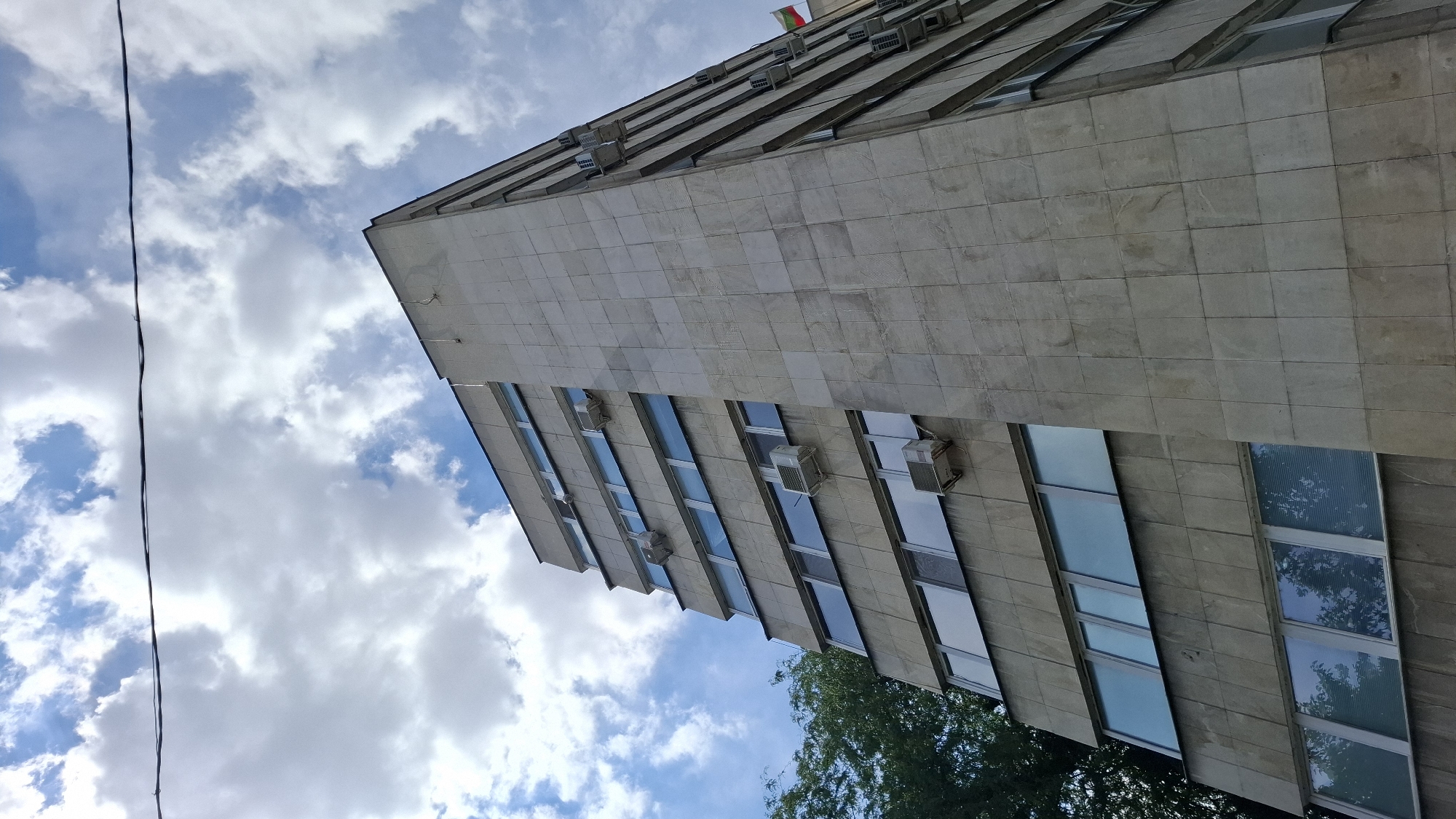 The image shows the exterior of a concrete building with a facade of large, light-colored square tiles. The building is viewed from a low angle, looking up towards the sky. Several rows of windows are visible, some with blue-tinted glass and others reflecting the cloudy sky and trees. Air conditioning units are mounted on the exterior of the building below many of the windows. A thin black wire runs vertically through the left side of the image. At the top of the building, a small flag with red, white, and green stripes is visible. The sky is filled with white, fluffy clouds against a blue background, suggesting daytime. Green trees are partially visible at the bottom of the frame. The overall scene appears to be an urban setting in Plovdiv, Bulgaria. No people are visible in the image, and no specific activity or event is taking place.