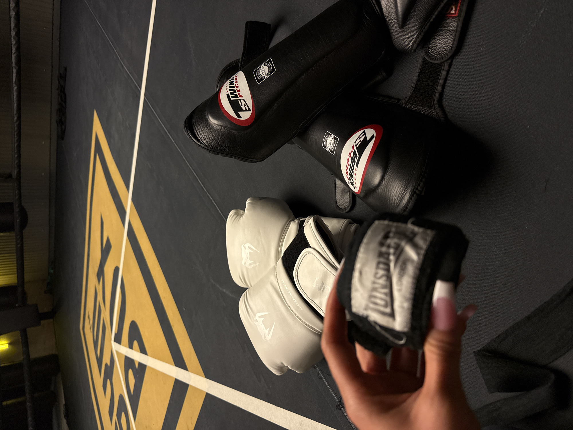 A close-up, eye-level shot captures boxing gear laid out on a black mat within a boxing ring. The mat is marked with yellow and white lines, forming a square pattern. In the foreground, a hand with pink-tipped nails holds a pair of black Lonsdale hand wraps. To the right, a pair of white Venum boxing gloves rests on the mat, and further back, two black Twins Special shin guards are positioned. The background reveals the edge of the boxing ring with dark ropes and a dimly lit area. The scene is likely within a gym or training facility in London, United Kingdom, given the presence of Lonsdale, a brand associated with the city.