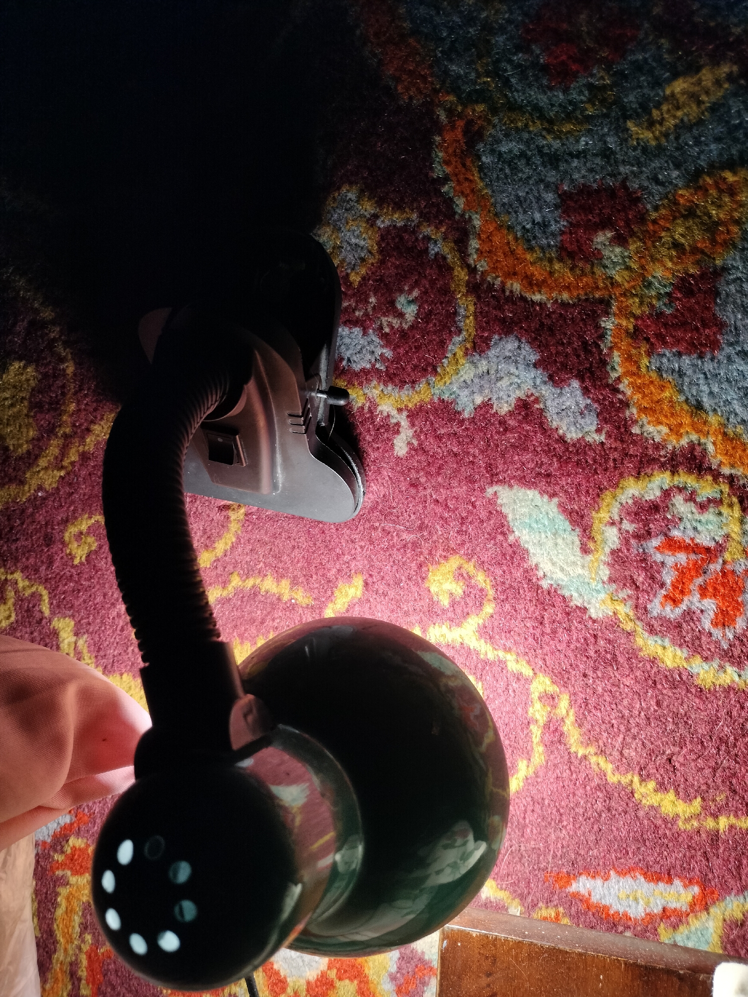 A close-up shot from a high angle shows a dark green desk lamp with a flexible gooseneck. The lamp is positioned on a carpet with a red, yellow, blue, and white intricate pattern. A hand, partially visible on the left, is holding the flexible neck of the lamp. The lamp head, which is dark green and has a circular design with several small, illuminated circles on its underside, is casting a shadow onto the carpet. The background is dark, with some of the carpet pattern visible. The scene is indoors, likely in a home or office environment, in Lykhove, Ukraine.