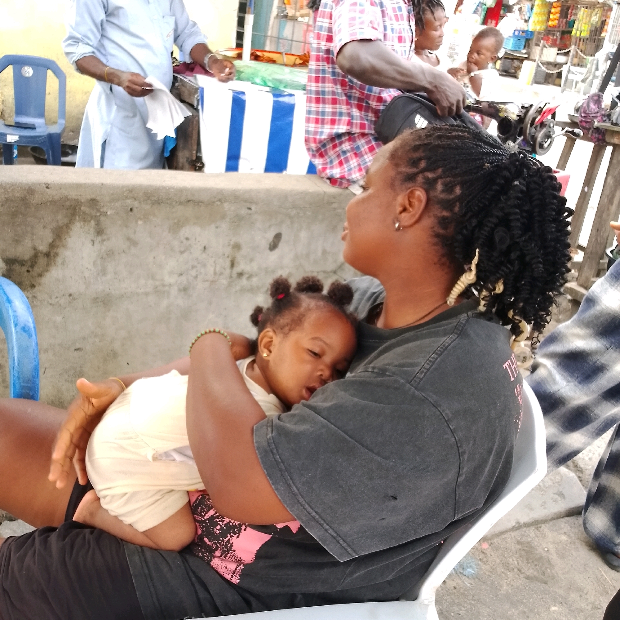 In Ado, Nigeria, a woman with dark skin and braided hair is seated, holding a sleeping toddler in her arms. The woman wears a dark t-shirt with faded lettering and a beaded bracelet on her left wrist. The toddler, also with dark skin, is dressed in light-colored clothing and rests its head on the woman's shoulder. The background shows a street scene with a concrete wall, a blue plastic chair, a sewing machine, and various market stalls, suggesting a commercial or residential area. Another person, partially visible, wears a plaid shirt. The overall environment appears to be outdoors, possibly in a market or roadside setting.