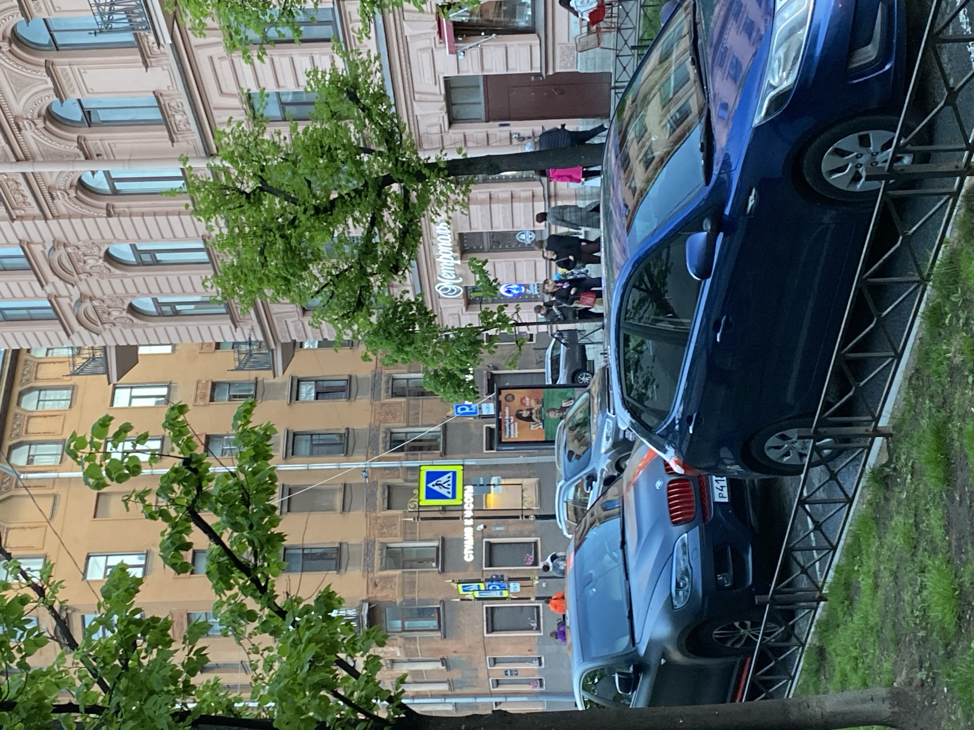 The image shows a street scene in Saint Petersburg, Russia. The foreground features two parked blue cars along the edge of a sidewalk with a metal railing. Behind the cars, a street is lined with buildings of varying heights and architectural styles. The buildings are primarily in shades of beige and brown, with multiple windows. Pedestrians are visible walking along the sidewalk in the distance. A green tree is partially obscuring the buildings and adds to the street's urban greenery. Street signs and advertisements are visible among the buildings, including a pedestrian crossing sign and a sign that reads "Memories" in Cyrillic.