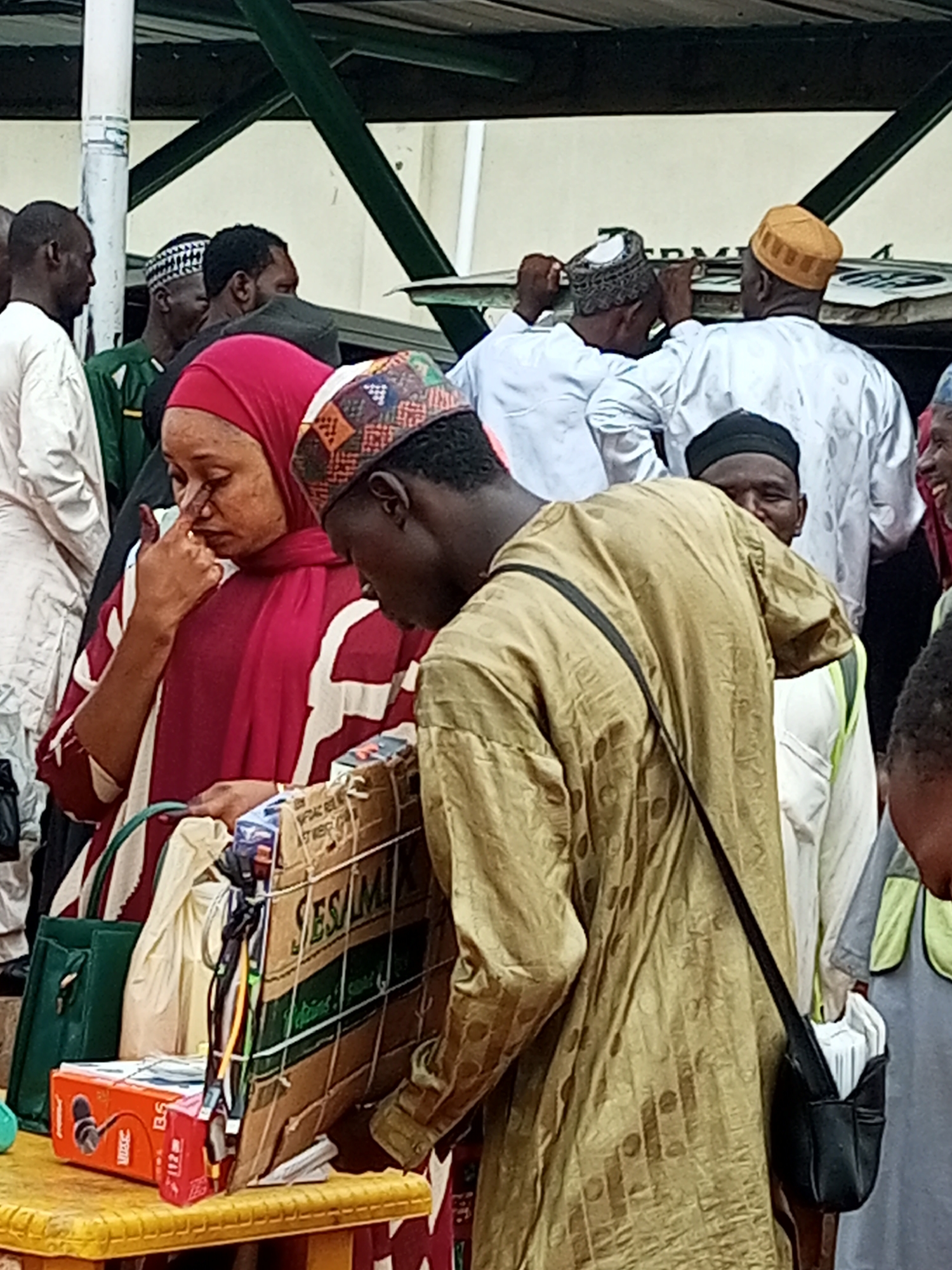 In Gombe, Nigeria, a group of people, some wearing traditional Islamic attire, are gathered outdoors, likely at a market or public square. The foreground features a woman in a red headscarf and patterned dress, her hand touching her face as if to wipe away tears or an itch. Beside her, a person in a gold-colored robe with a patterned design on the head covering is looking down towards a display of goods on a yellow table. These goods include what appear to be electronic accessories, with a red box labeled "BS" and a larger cardboard box containing various items. In the background, several men are visible, some wearing white robes and caps, suggesting a religious or cultural gathering. The environment appears somewhat crowded and bustling, typical of a Nigerian marketplace.
