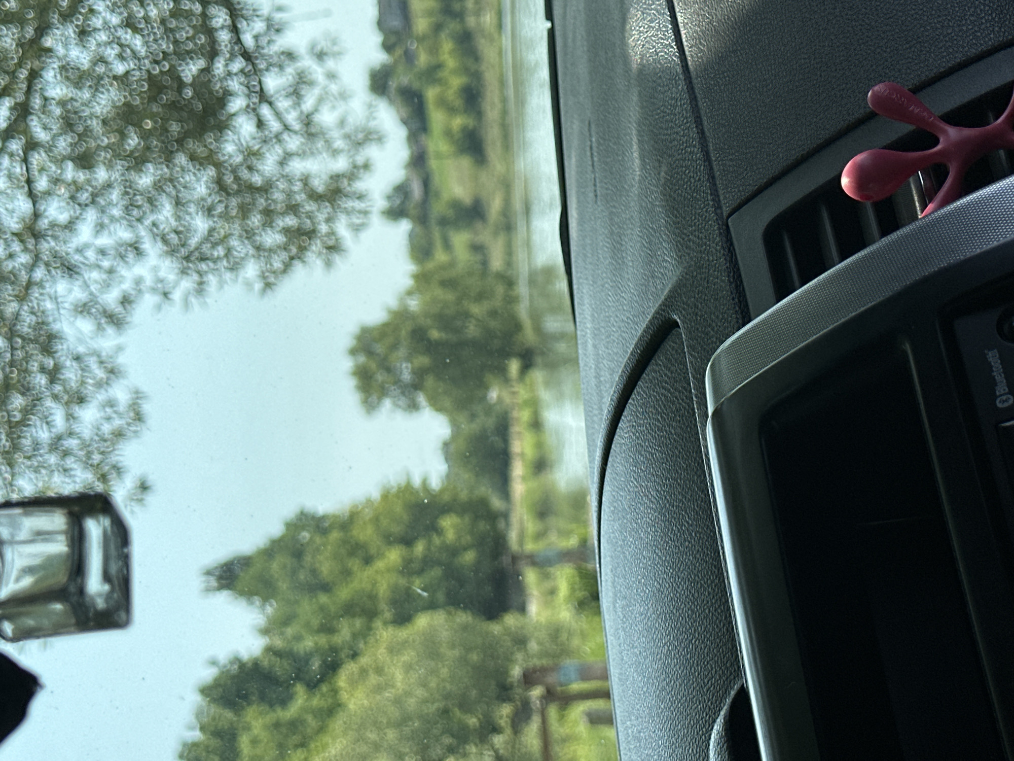 This is a photograph taken from inside a vehicle, looking out through the windshield. The view is of a body of water, likely a river or lake, bordered by lush green trees and vegetation. In the background, several houses are visible, suggesting a rural or semi-rural setting. A faint reflection of a car's rearview mirror is visible at the top of the windshield, and a small, red, octopus-like air freshener is attached to the car's vent on the right side of the dashboard. The lighting suggests it is daytime, with an overcast or hazy sky. The location is identified as Makiv, Ukraine.
