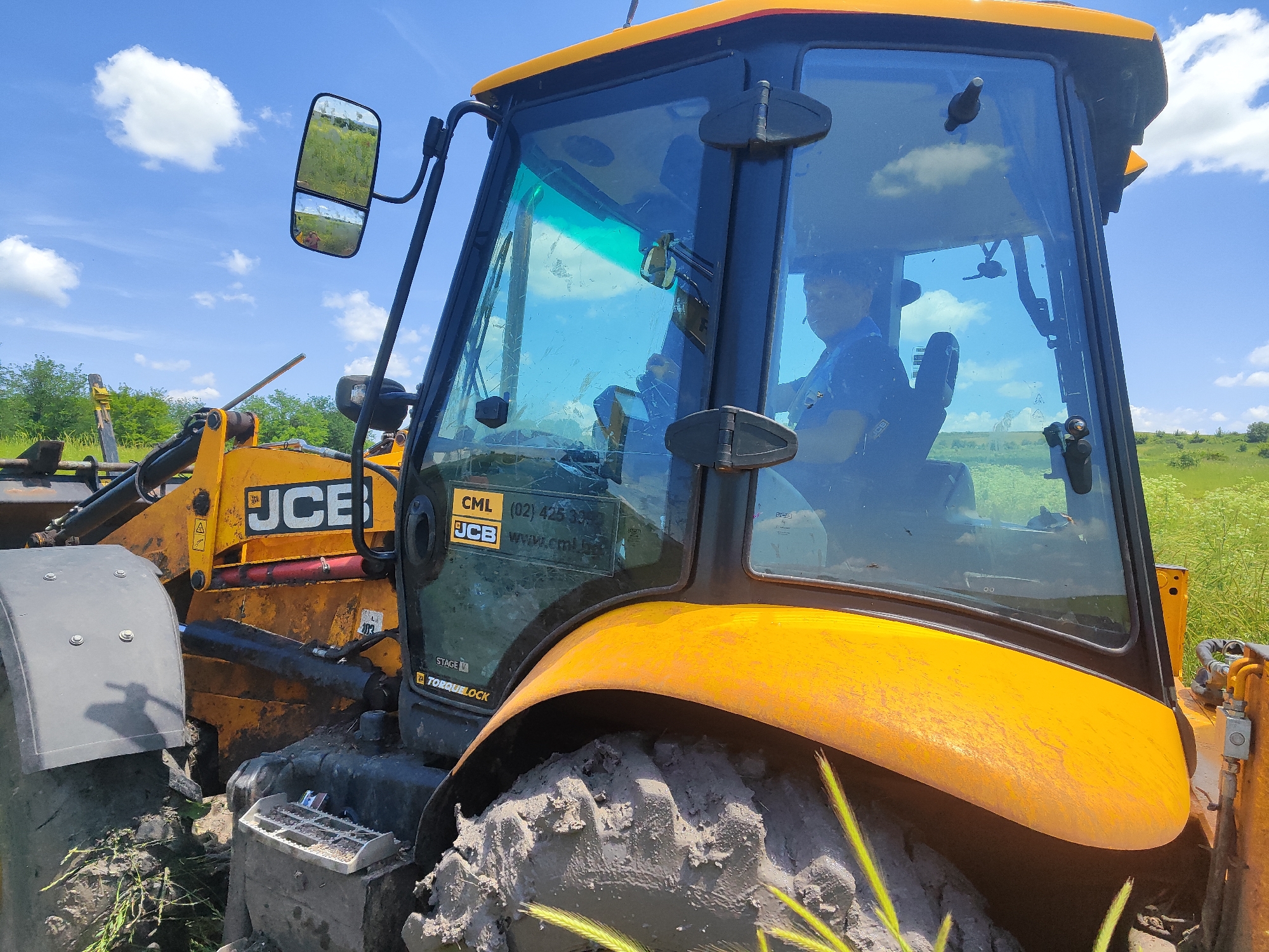 A yellow JCB backhoe loader is parked on a muddy field under a bright blue sky with scattered white clouds. A person wearing a blue shirt and dark cap is seated in the driver's seat, looking forward. The backhoe has large tires, and the cabin windows are visible, reflecting the surrounding environment. The machine is partially obscured by mud and grass. The scene appears to be in Malak Manastir, Bulgaria, based on the context provided.
