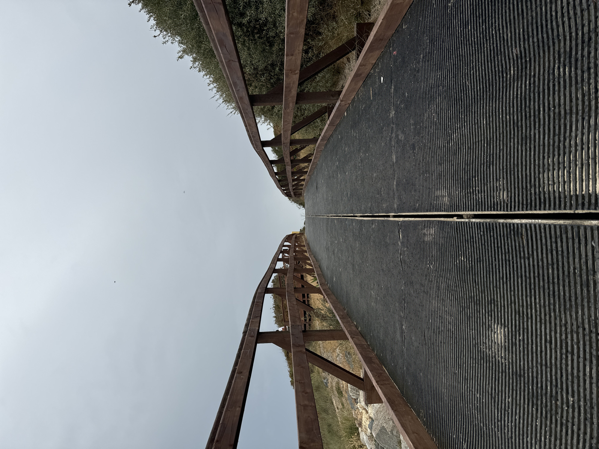 A wooden footbridge with dark, ribbed decking curves upwards into an overcast sky in Courtown, Ireland. The bridge is bordered by wooden railings and traverses a natural landscape with rocks and vegetation. The sky is a pale, uniform gray, with a small dark object, possibly a bird, visible in the upper left quadrant. The overall scene is one of a utilitarian structure integrated into a coastal environment.