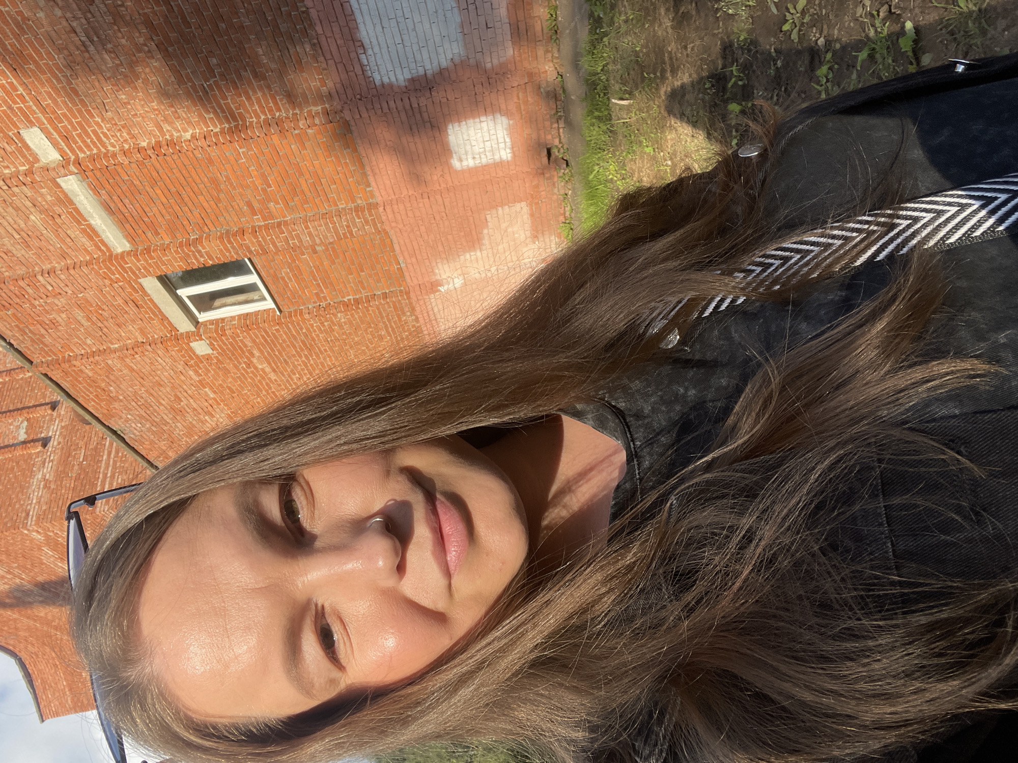 A woman with long brown hair, wearing sunglasses on her head and a nose ring, smiles at the camera. She is wearing a dark-colored denim jacket over a dark-colored top. A patterned strap is visible across her shoulder. She is standing outdoors in front of a red brick building. The building has a white-framed window and some light-colored rectangular patches on the brickwork. The ground in front of the building is covered with dirt and sparse green grass. The image appears to be taken during the daytime, with sunlight casting shadows. The woman's location is confirmed to be Izhevsk, Russia.