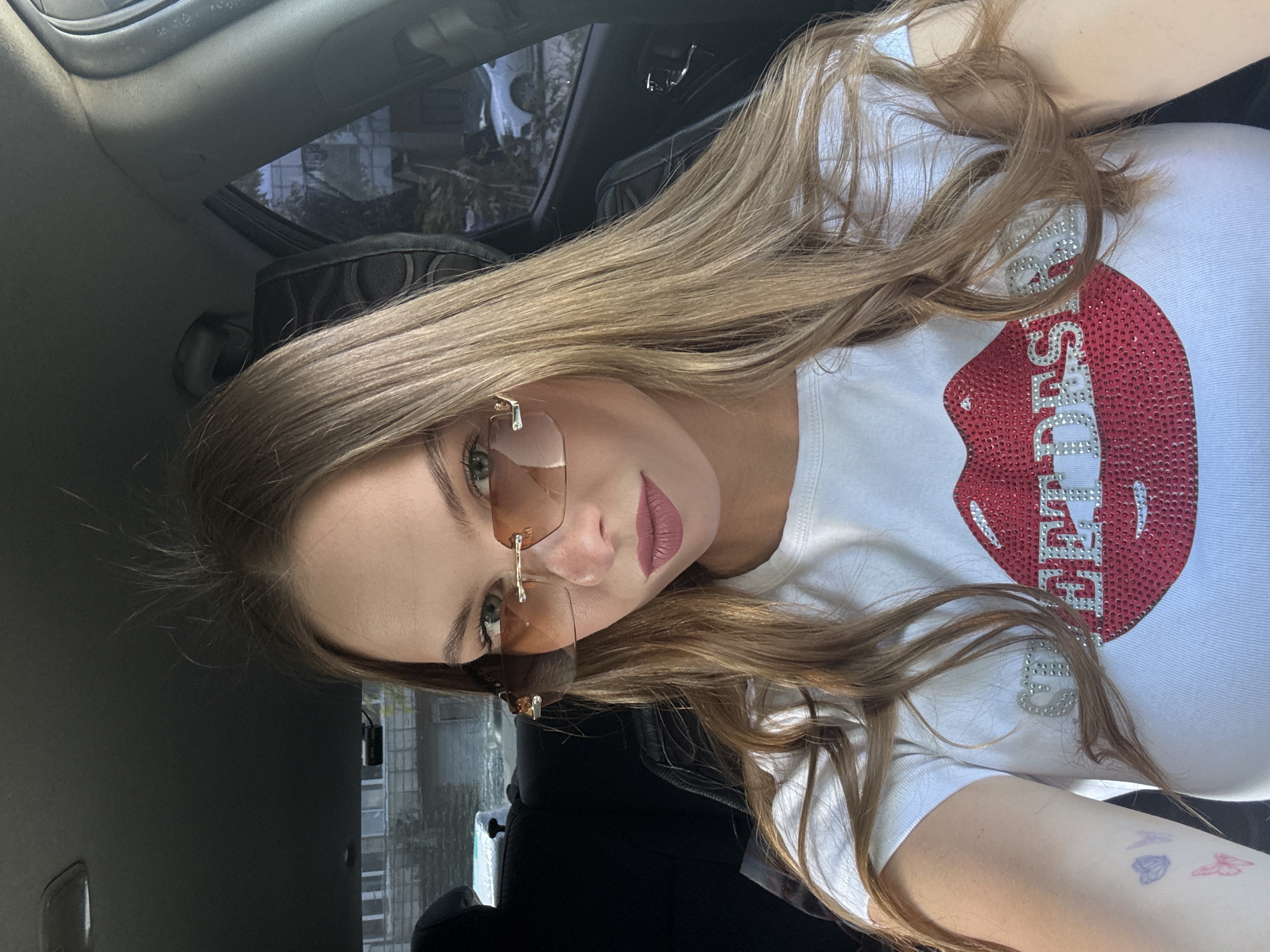 A young woman with blonde hair and green eyes wears sunglasses and a white t-shirt with a rhinestone lip graphic and the words "Street Desire" also in rhinestones. She is sitting in the driver's seat of a car, with the interior visible. The background shows buildings and trees, suggesting an urban setting. This image was taken in Perm, Russia.