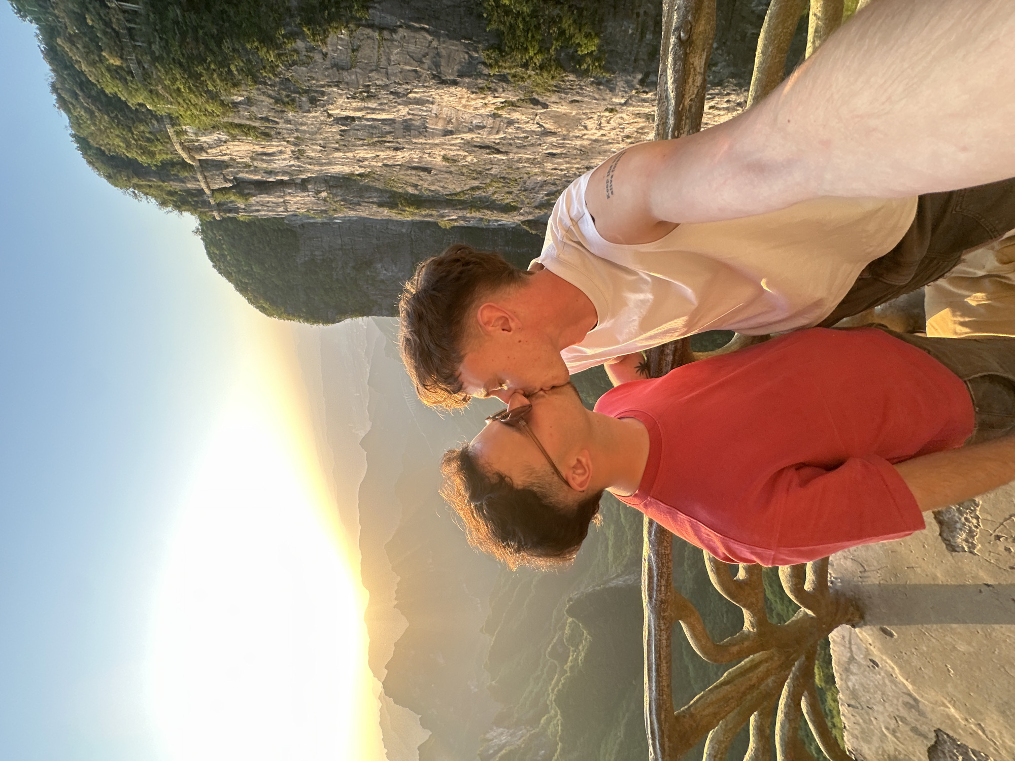 Two men are kissing against a mountain background in Yongding District, China. The man on the left is wearing a white t-shirt, and the man on the right is wearing a red t-shirt and eyeglasses. They are standing behind a decorative railing. In the background is a mountain and a bright sky.