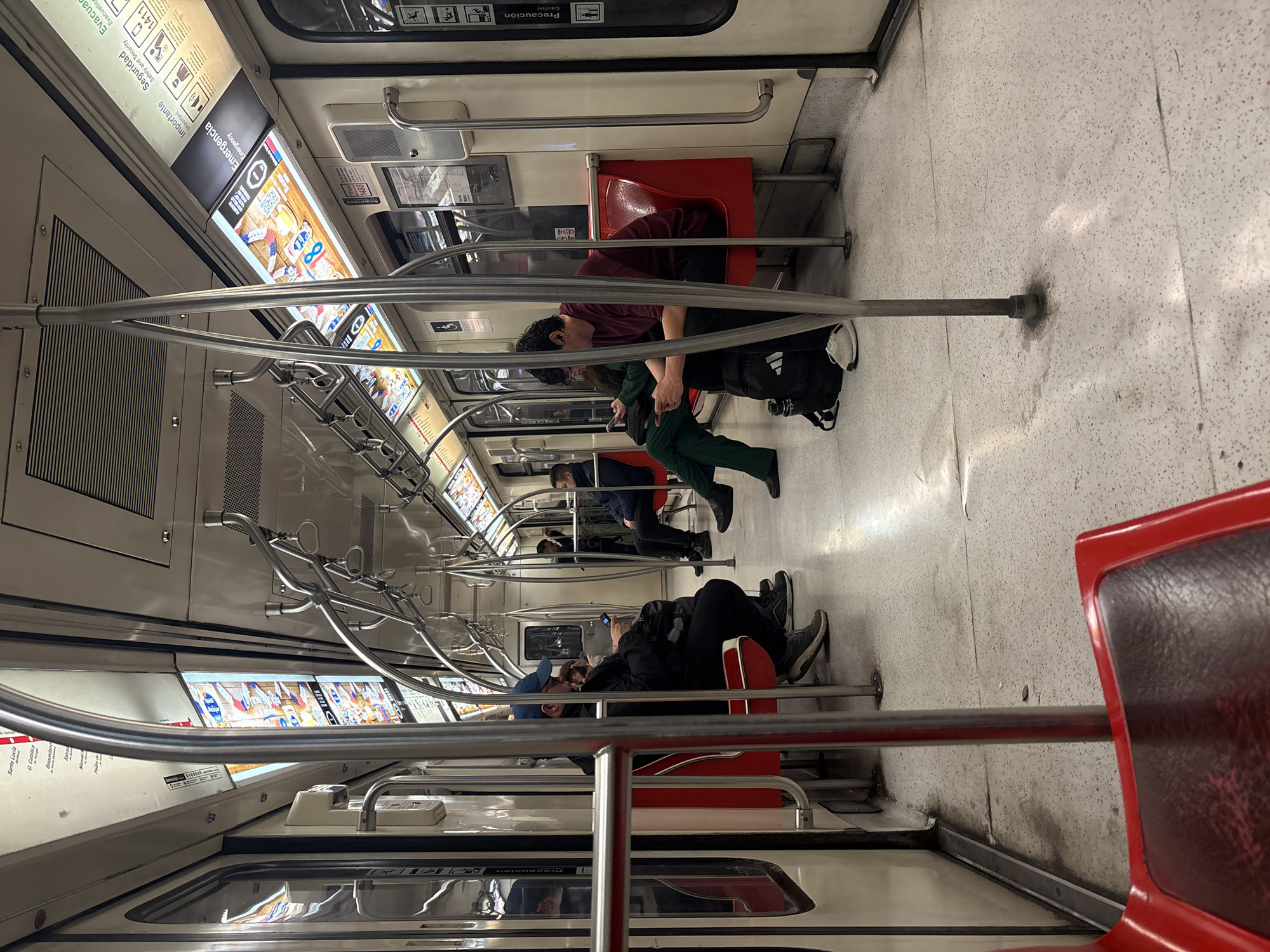 The image depicts the interior of a subway car, likely in Santiago, Chile, based on visible signage. Several passengers are seated on the red and white upholstered seats. On the left, a person wearing a blue cap and black jacket is looking at their phone. To their right, another passenger in a dark jacket is also seated. In the center and to the right, a person with curly hair, wearing a maroon t-shirt and black pants, is looking at their phone, with a black backpack at their feet. Further back on the right, another passenger in a dark jacket is seated. The subway car features chrome handrails and poles for standing passengers, and illuminated advertising panels are displayed above the seats. The floor is a light-colored speckled material, showing some discoloration and dampness in places. The overall environment appears to be a typical urban transit setting.