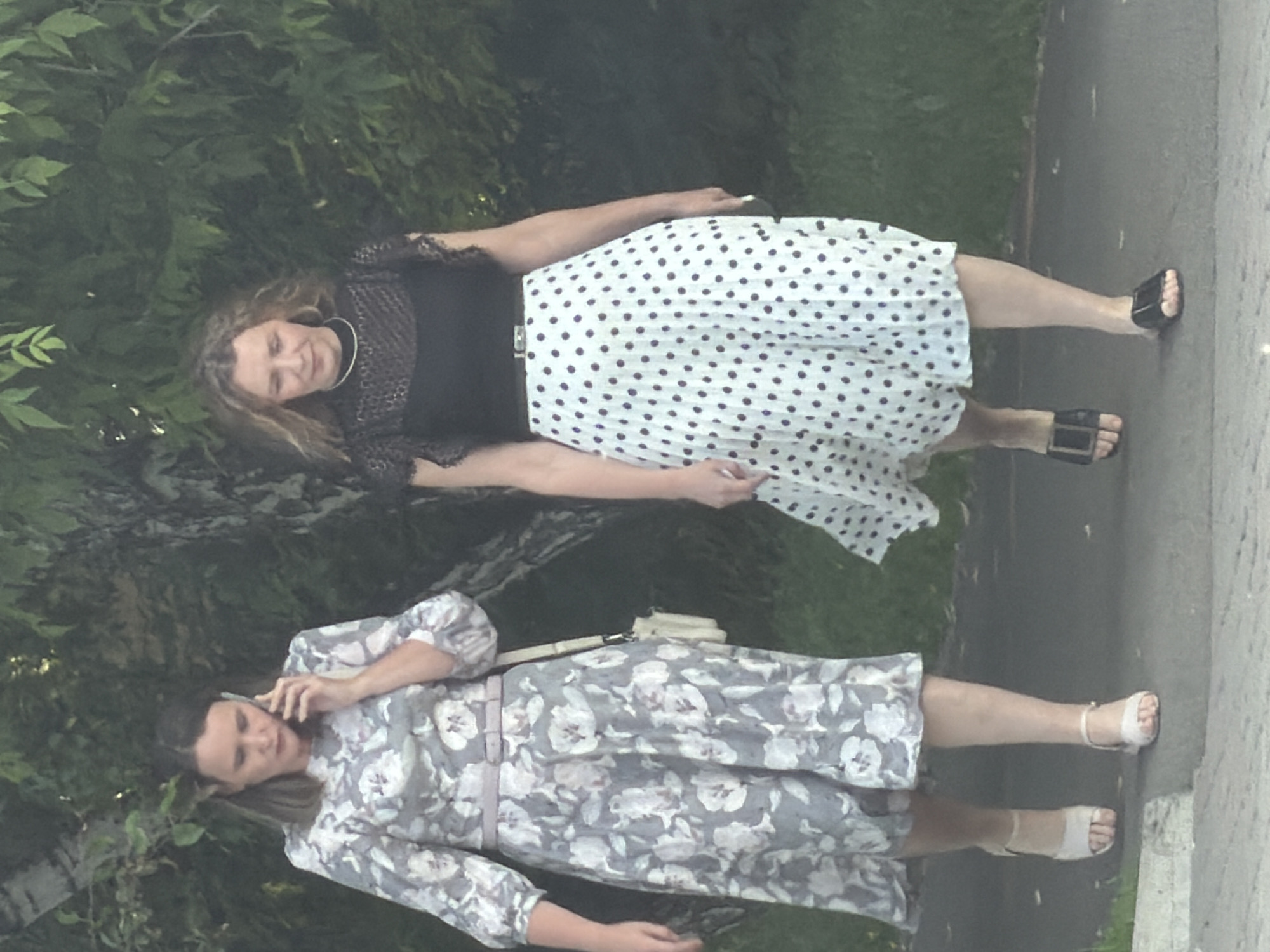 Two women are walking on a paved path outdoors. The woman on the left is wearing a grey floral dress and white sandals. She is holding a cell phone to her ear. The woman on the right is wearing a black lace top and a white polka dot skirt with black polka dots. She is wearing black sandals. The background consists of trees and foliage, suggesting a park or a residential area. The location is the city of Krasnoyarsk, Russia.