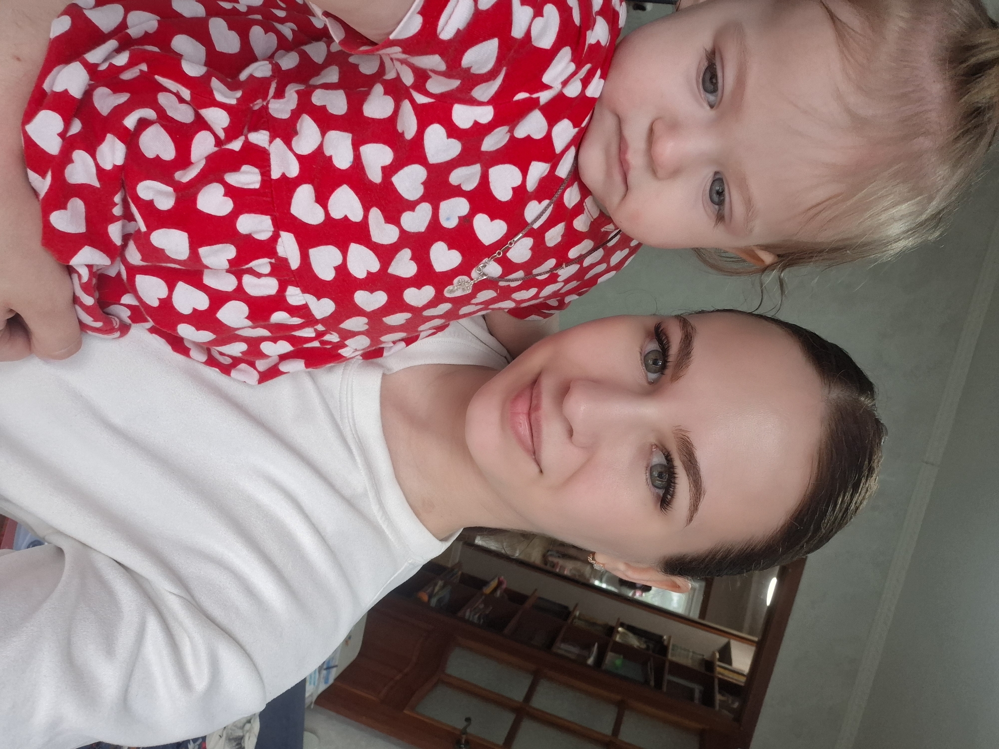 A selfie shows a young woman holding a baby. The woman has her hair pulled back and is wearing a white long-sleeved shirt. The baby is wearing a red outfit with white hearts. The location is in a room with a wooden cabinet in the background. It is plausible that the image was taken in Chaikovsky, Russia.