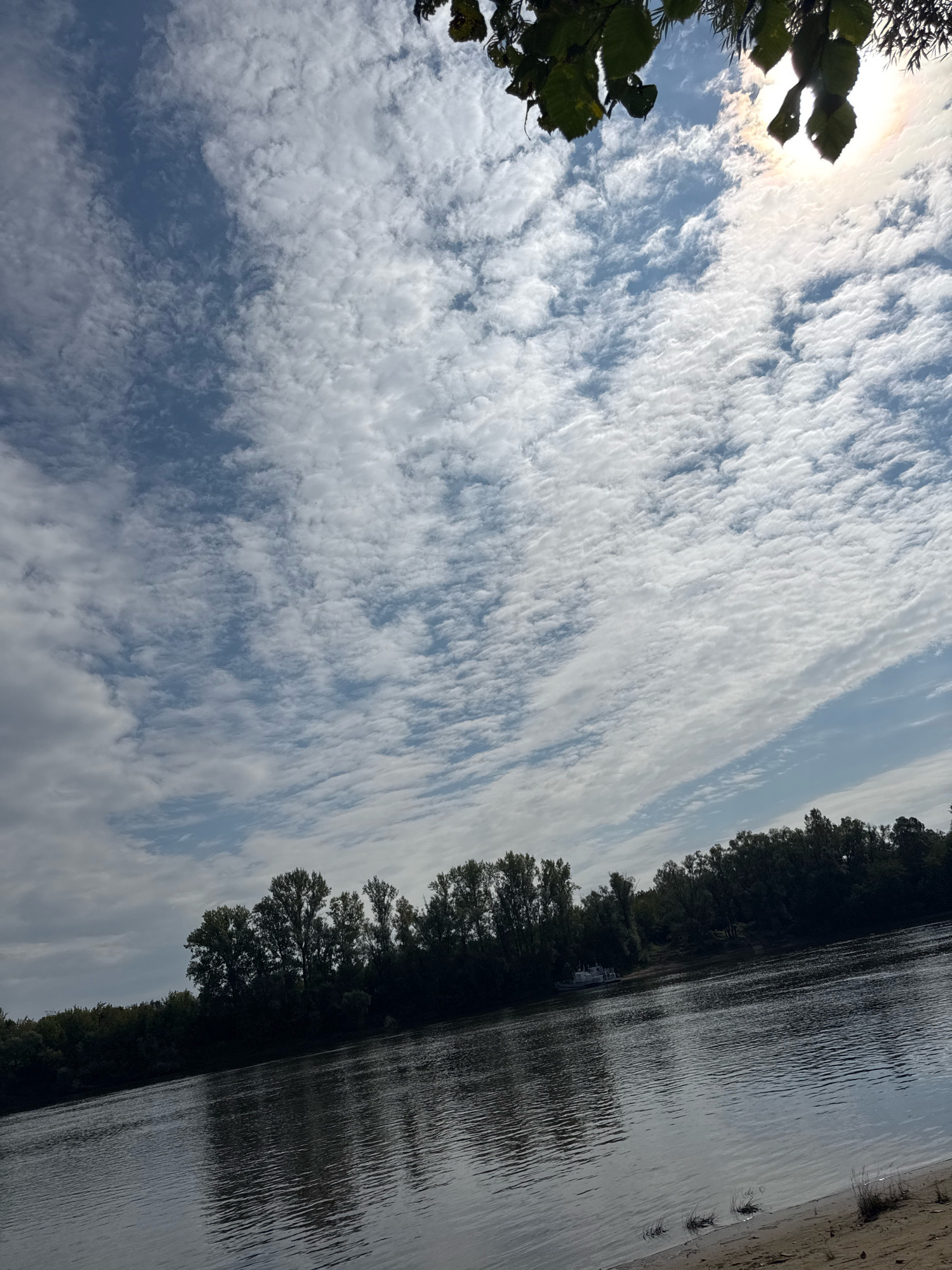 The image depicts a wide river with a sandy bank in the foreground. A dense line of trees forms the opposite bank, casting reflections on the water's surface. A small white boat is visible near the shore on the far side. The sky is filled with scattered white clouds against a bright blue background, with the sun partially obscured by foliage in the upper right corner, creating a lens flare. This scene is likely located along the Belaya River in Ufa, Russia, a common location for such landscapes. There are no people or animals visible in the image.