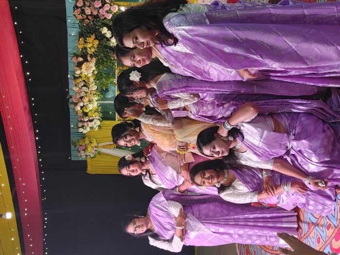 A group of eight women in lavender sarees are arranged in a staggered line, with flowers decorating the wall behind them. The sarees appear to be made of a shiny material. The women are all looking in different directions, and the lighting is soft. One woman in the center wears a pale yellow or gold saree. A hand is visible in the lower right corner. The overall impression is of a festive event or celebration.