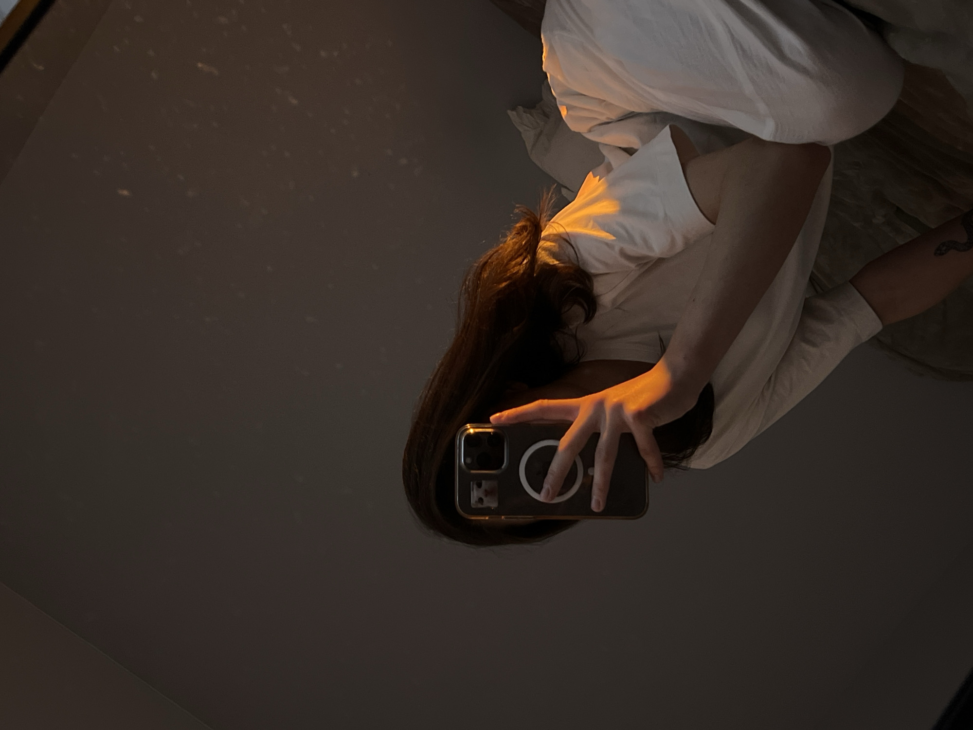 The scene depicts an individual indoors in Ternopil, Ukraine, under low light conditions. A person with dark hair is seated or reclining on a bed, partially covered by white sheets and pillows, and wearing a light-colored short-sleeved t-shirt. The individual holds a dark smartphone, fitted with a clear case featuring a prominent MagSafe ring, in their right hand. The phone's orientation obscures the face, but a small, light-colored, cat-shaped image is discernible on the phone's display area via reflection. A snake-like tattoo is present on the left forearm. A directed orange-yellow light source illuminates the individual's hair, right shoulder, right hand, and sections of the bedding. This light sharply contrasts with the dark, unadorned background wall. Faint specks, consistent with dust particles or minor reflections, are visible on the upper right section of the wall. A subtle diagonal line on the upper left wall suggests a structural joint. The posture indicates the individual is taking a mirror self-portrait.
