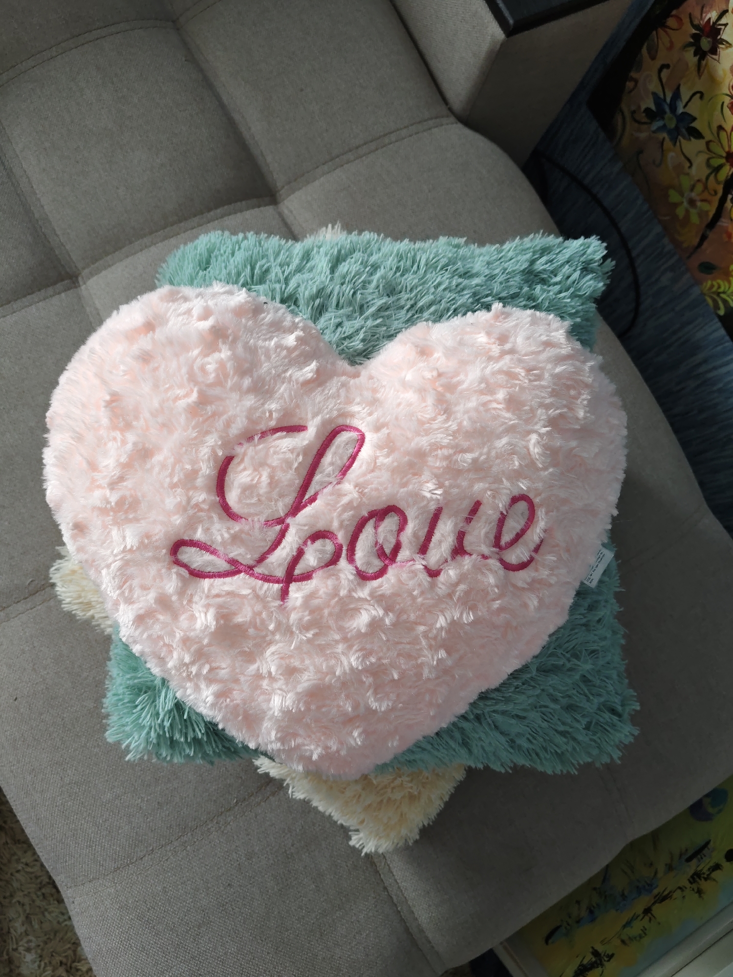 The image shows a close-up, overhead view of a pink heart-shaped pillow with "Love" embroidered in dark pink thread. The pillow is resting on a teal-colored, fluffy pillow. Below the teal pillow is a cream-colored fluffy pillow. These pillows are placed on a gray upholstered surface, possibly a sofa or chair, which has a quilted pattern. The background is blurred, but appears to include a dark blue fabric with a floral pattern. There are no people visible in the image. The scene is indoors. The image was taken in Rostov-on-Don, Russia.