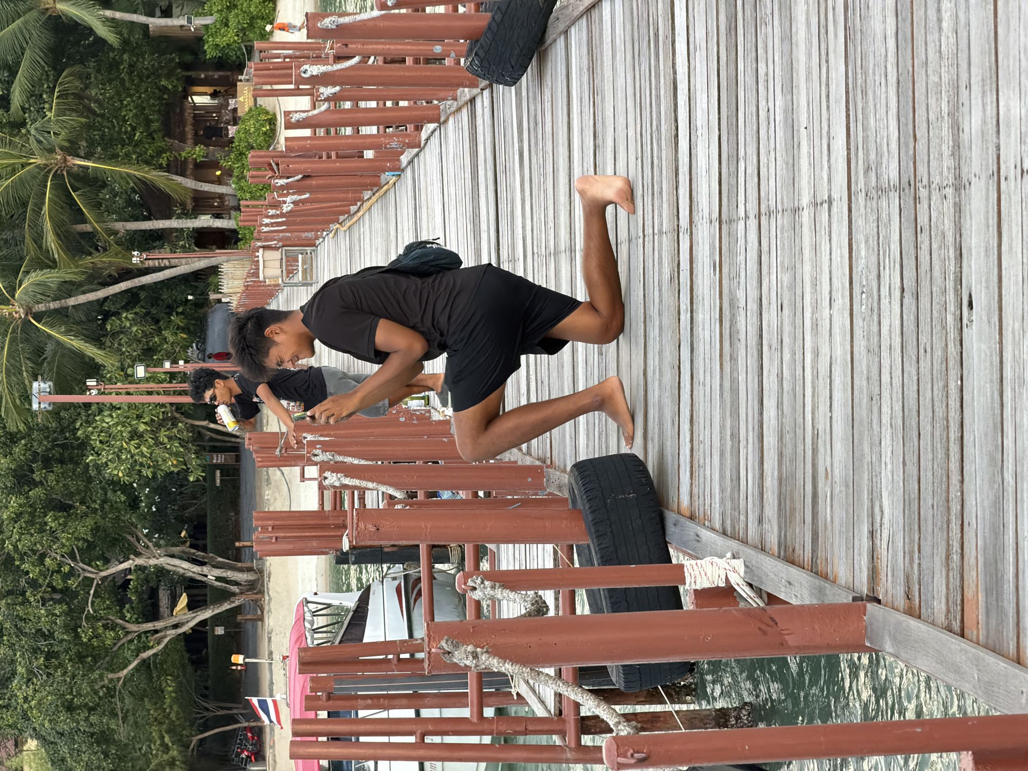 The image depicts a wooden pier with a person in the foreground performing a dynamic movement, possibly a jump or a split. They are wearing a black t-shirt and black shorts, and their body is angled as they crouch down with one leg extended. Barefoot, they appear focused on their action. Another person is visible further down the pier, partially obscured and facing away, seemingly engaged with something on the pier.
The pier is constructed of weathered wooden planks and is lined with reddish-brown railings. Ropes are secured to these railings, suggesting a functional docking area. A discarded tire is visible on the pier, likely used as a bumper. In the background, lush green palm trees and dense foliage suggest a tropical environment. A glimpse of buildings with thatched roofs can be seen among the trees, with some people milling about. The water below the pier is calm and reflects the surroundings. Boats are docked along the pier, one with a prominent pink canopy. A Thai flag is visible on a pole near the boats.
The scene appears to be set during the day, possibly late afternoon given the soft lighting and shadows. The overall mood is relaxed and casual, typical of a tropical vacation spot. The location cues such as palm trees, the Thai flag, and the style of the boats strongly indicate Baan Bang Rak, Thailand. No discernible text is visible in the image beyond what might be on a distant sign.