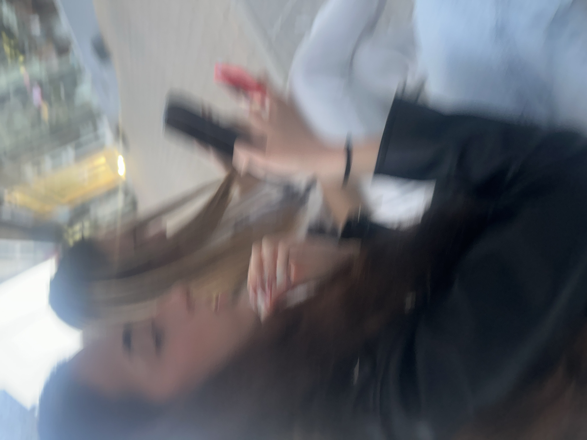 The image shows two young women, out of focus, with one on the left and the other on the right. The woman on the left has dark hair and is wearing a black jacket. She is holding a phone in her right hand and appears to be looking at it. The woman on the right has lighter hair and is wearing white pants and a light-colored top. She is holding a red phone in her right hand and a black object, possibly another phone, in her left hand. They are seated next to each other, with a blurred background suggesting an outdoor urban setting, potentially in Shumen, Bulgaria, given the prompt. The lighting suggests it is daytime.