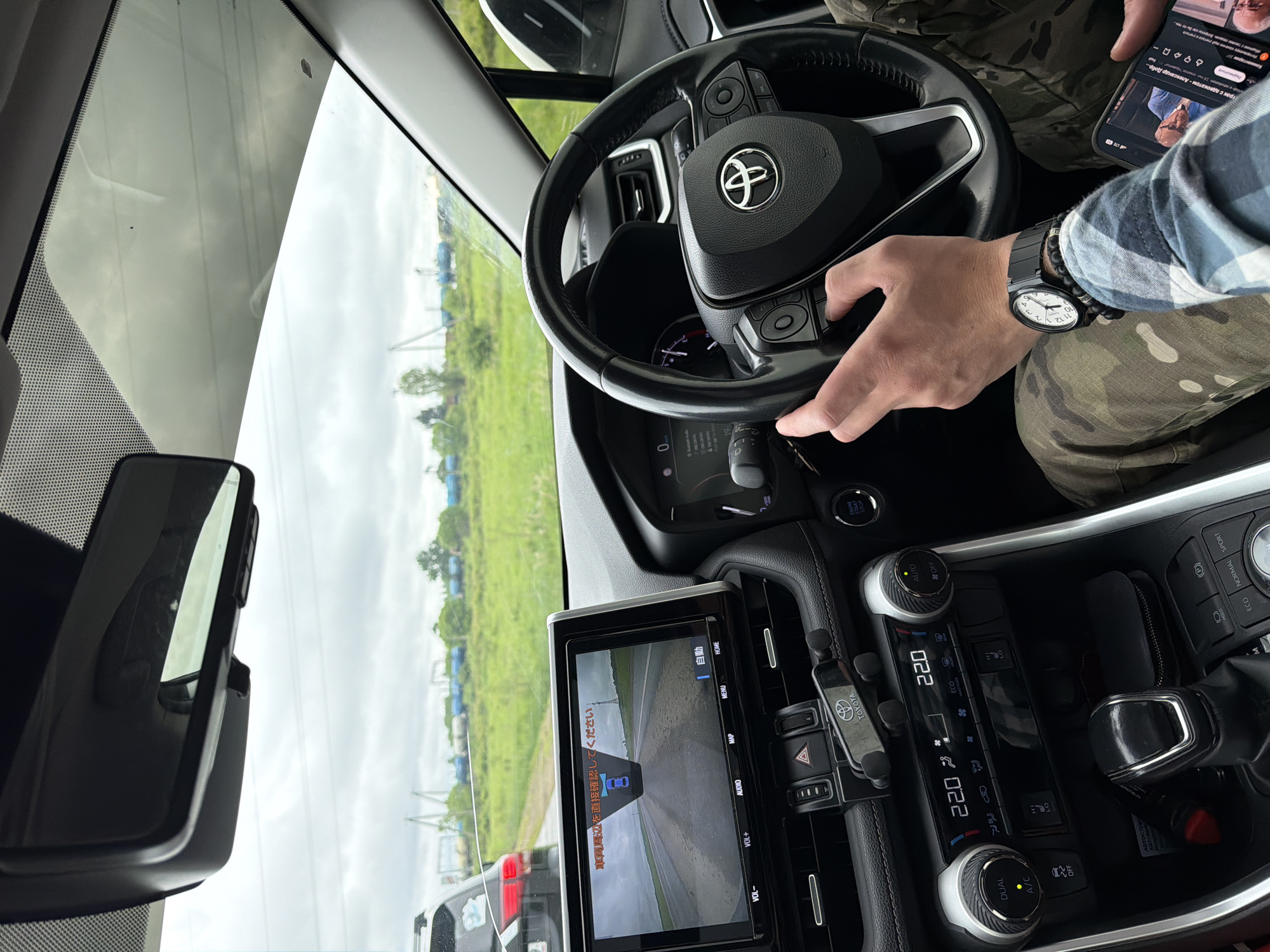 The image shows the interior of a car, likely a Toyota RAV4, viewed from the passenger's perspective. A person's hands are visible on the steering wheel and holding a smartphone. The car's dashboard displays various controls and a central infotainment screen showing a rear-view camera feed with Japanese text. Outside the car, a rural landscape with green fields and cloudy sky is visible. The location is identified as Kirga, Russia.