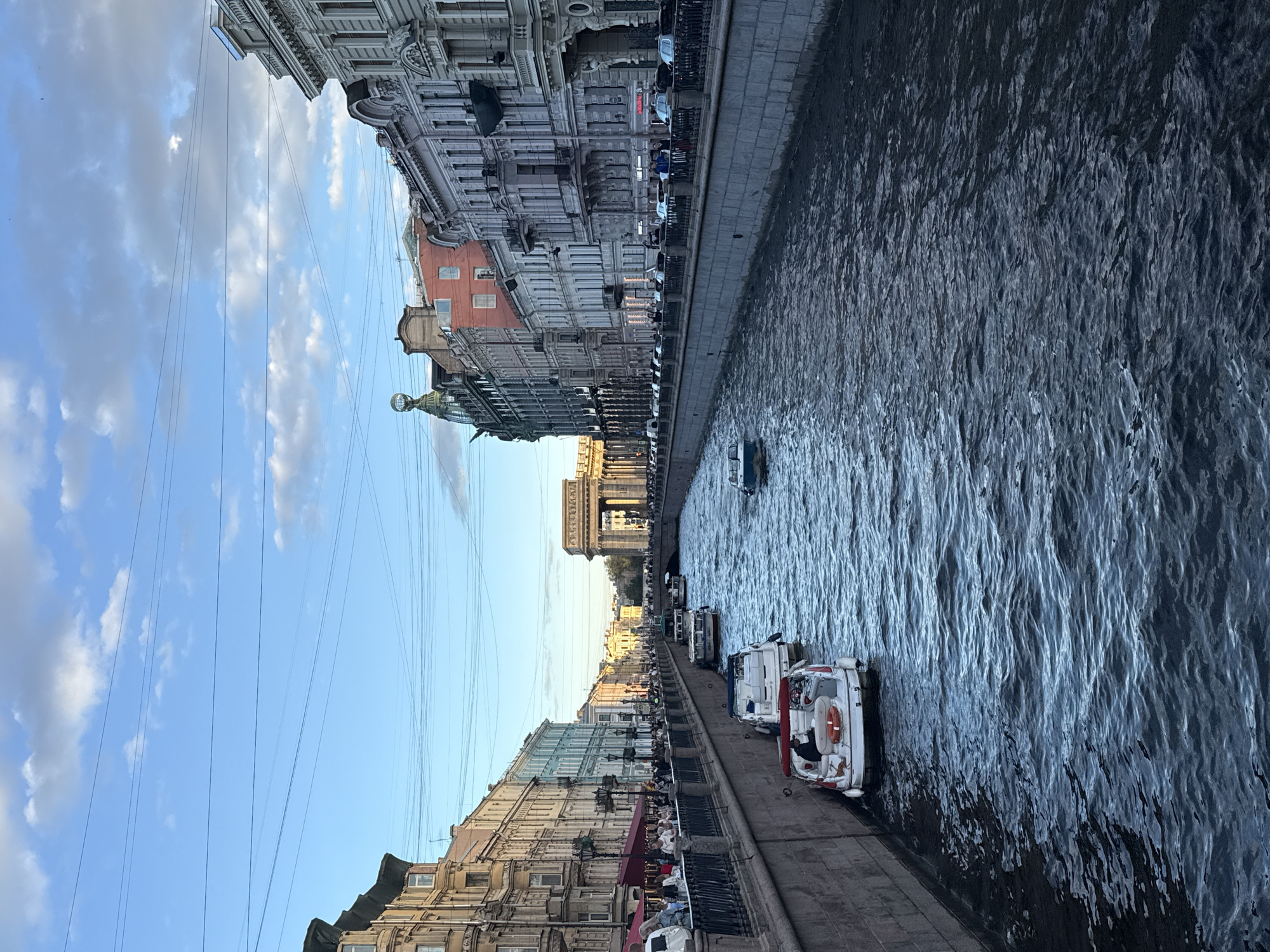 A vertical, eye-level shot captures a sunlit canal in Saint Petersburg, Russia. The canal, bordered by stone embankments, flows into the distance towards a grand archway. Several boats, including two white motorboats with red accents, are moored or cruising on the water. The right embankment is lined with ornate, multi-story buildings featuring detailed facades and balconies. The left embankment shows a row of buildings in varying architectural styles and colors, with a pedestrian walkway populated by numerous people. The sky above is a clear blue with scattered white clouds, and power lines crisscross the upper portion of the frame. The sunlight casts a warm glow on the scene, highlighting the architectural details and the texture of the water.