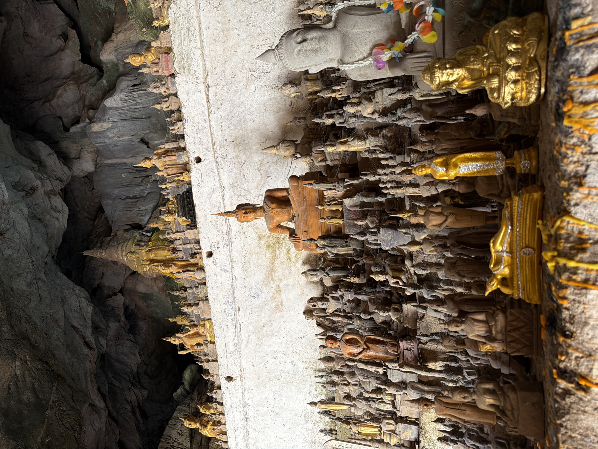 The image depicts the interior of a cave in Muangkeo, Laos, filled with numerous Buddha statues of varying sizes and materials. The statues are arranged on tiered shelves along the cave wall and in the foreground. The cave ceiling is rough and rocky, with stalactites visible. In the foreground, there are several prominent golden Buddha statues, including one in a reclining posture and another seated. A larger, grey stone Buddha statue adorned with a garland of colorful flowers is visible to the right. The overall scene is one of religious devotion and art within a natural cavern.