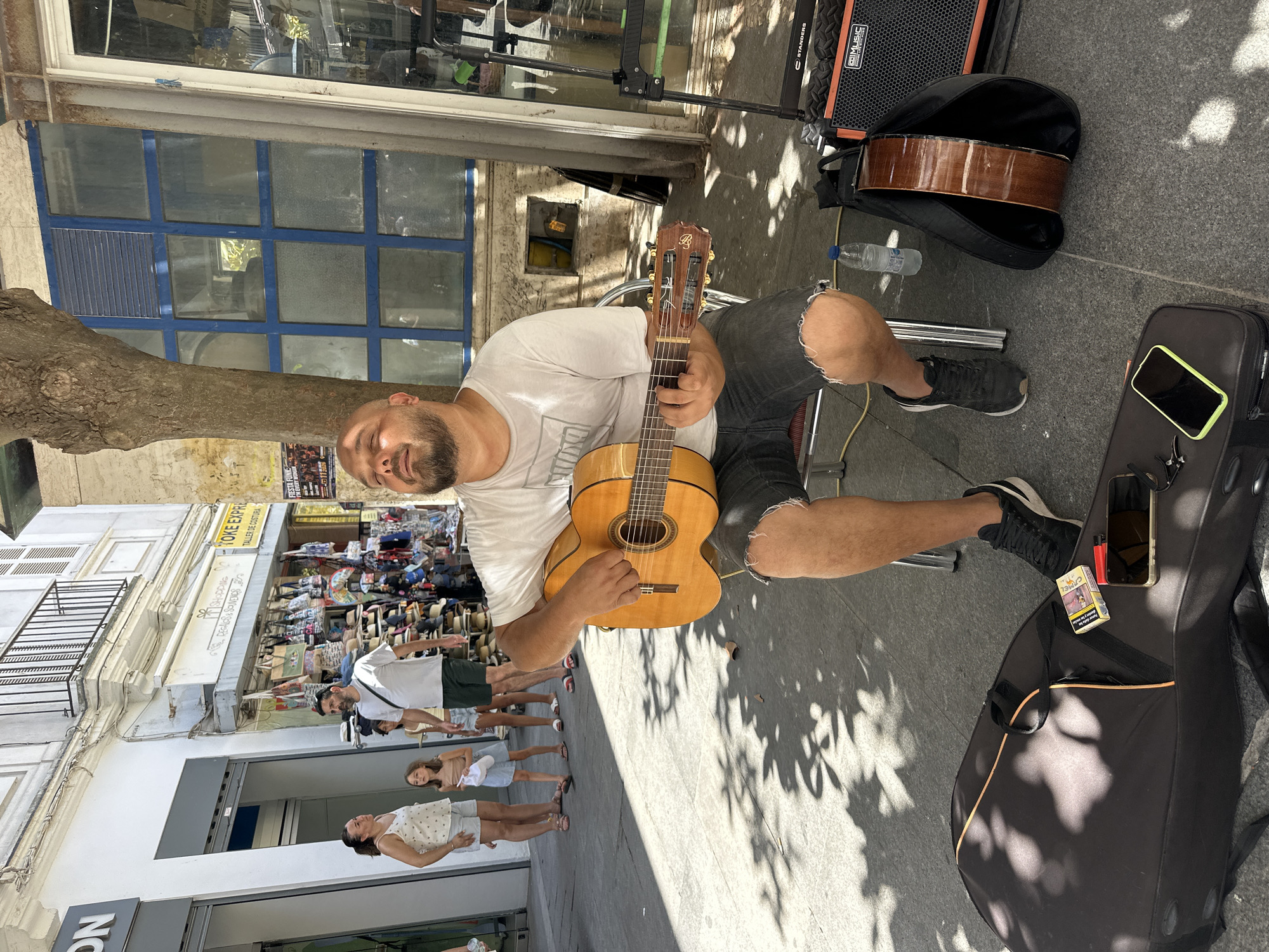 A man sits on a folding chair on a paved street in Cádiz, Spain, playing a classical guitar. He wears a white t-shirt and ripped denim shorts, with black sneakers. His eyes are closed, and his head is tilted back, appearing relaxed or lost in his music. In front of him, on the ground, is a black guitar case, a pack of cigarettes, a lighter, and two smartphones. To his right, another guitar is partially visible in a black gig bag next to a small orange and black amplifier. In the background, several people are walking on the sidewalk in front of shops, including a woman with a ponytail, a young girl, and a man in a white t-shirt and dark shorts. The street is shaded by a tree, casting dappled shadows on the ground. The architecture suggests a European city, and the signage in the background indicates shops selling souvenirs and clothing.