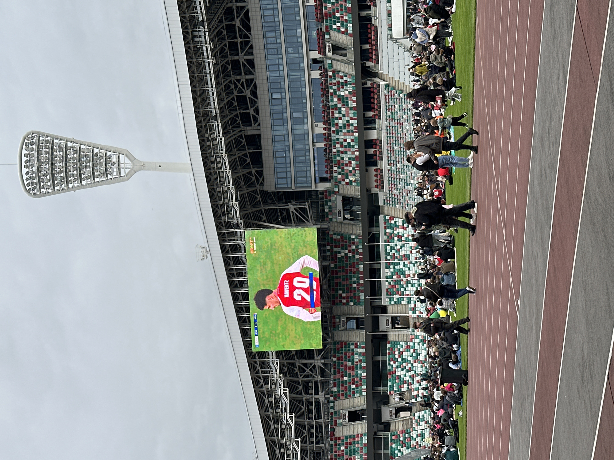 The image captures a scene at the Dinamo National Olympic Stadium in Minsk, Belarus. A large video screen displays a soccer player wearing a red and white jersey with the number 20. Stadium seating is visible with a mix of green, red, and white colors. Spectators can be seen in the stands and walking along the running track adjacent to the field. A stadium light fixture is positioned above, and the sky appears overcast. The running track itself shows multiple lanes with varying shades of red and gray.