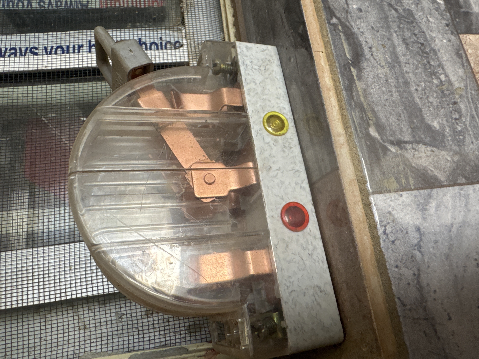 The image shows a close-up of a round, clear plastic electrical switch with copper internal components. The switch has a gray handle sticking out from the top. Attached to the side of the switch is a white rectangular piece with two circular indicators: a yellow one near the top and a red one below it. The switch is mounted on a surface, and behind it, a mesh screen is visible, suggesting it might be part of a window or door screen.
In the background, there's a glimpse of what appears to be text on a white surface behind the mesh screen. Part of the text is legible, reading "vays your hoice". The surrounding environment appears to be an indoor setting, possibly a home or a building under construction or repair, given the presence of unfinished surfaces and tiling visible to the right of the switch. The lighting suggests it's daytime. The overall impression is of an electrical component in a functional, perhaps slightly utilitarian, setting within a residential or commercial space in Abuja, Nigeria. There are no people visible in the image.