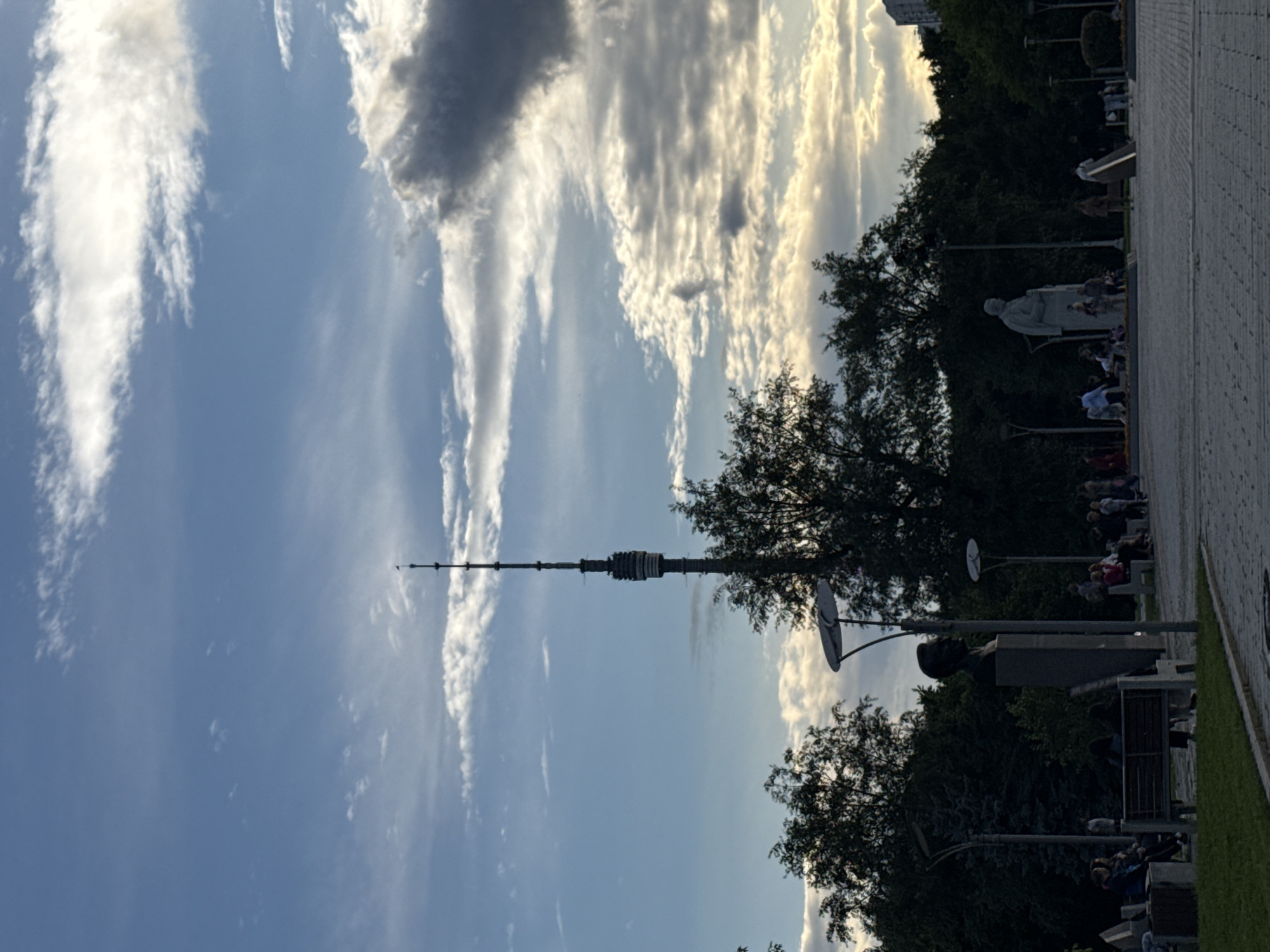 The image depicts a park scene in Moscow, Russia, with the Ostankino Tower prominently visible in the background. The tower, a tall, slender structure with a spire, stands against a sky with scattered clouds, some of which are dark and dramatic. In the foreground, several people are seated on benches and standing in small groups, engaged in leisure activities. There are statues in the park, including a bust and a larger, standing figure. Trees line the background of the park, with some buildings visible in the far distance. The overall impression is of a public space being utilized for recreation and relaxation.