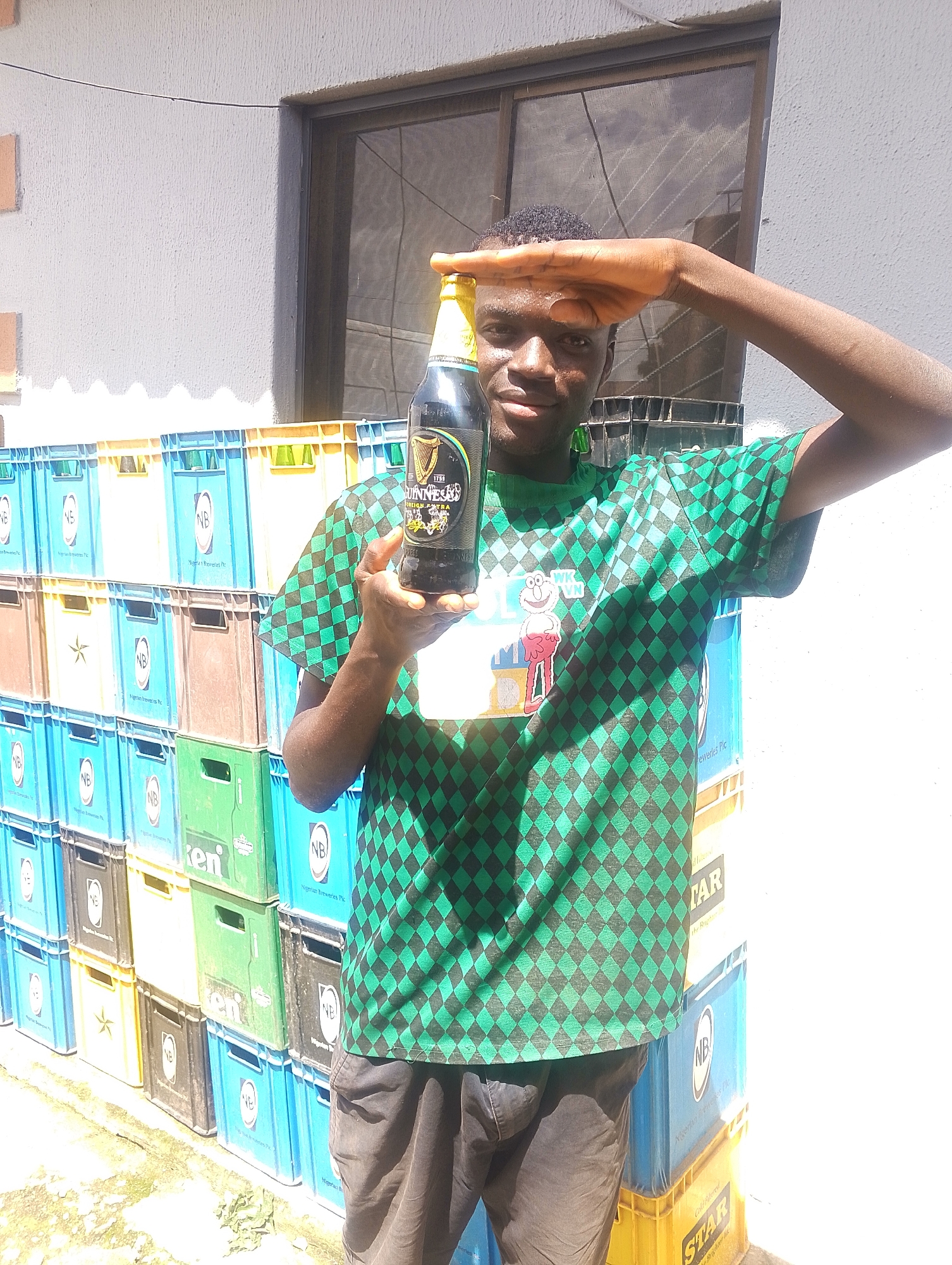 In this medium shot, a young Black man stands in front of a wall of stacked crates, possibly in an outdoor storage area in Jos, Nigeria. He is holding a bottle of Guinness Foreign Extra Stout in his right hand and has his left hand raised to his forehead as if shielding his eyes from the sun. The man is wearing a green and black checkered t-shirt with a cartoon graphic of Elmo on the front, and dark trousers. The crates are a mix of colors, including blue, green, yellow, and brown, and many are marked with logos and text such as "NB" for Nigerian Breweries Plc. The lighting suggests it is daytime, with bright sunlight casting strong shadows. The man's gesture and the bright light imply a sunny day.
