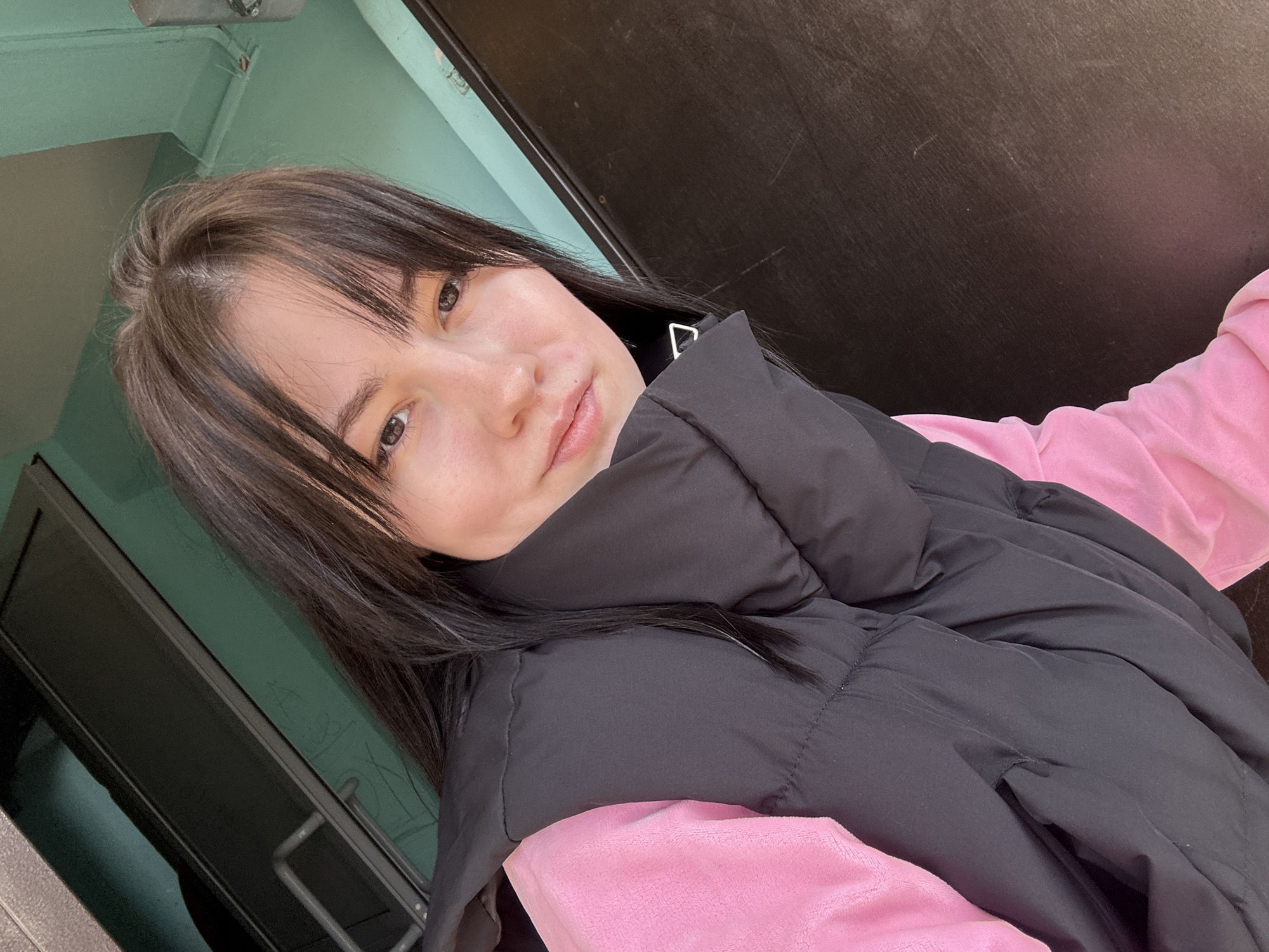 A young woman with brown hair and a pink sweater is wearing a black puffer vest. She is indoors, likely in the city of Chaikovsky, Russia. The background features a green wall and a dark door frame. The lighting is bright, and the photo appears to be a selfie.