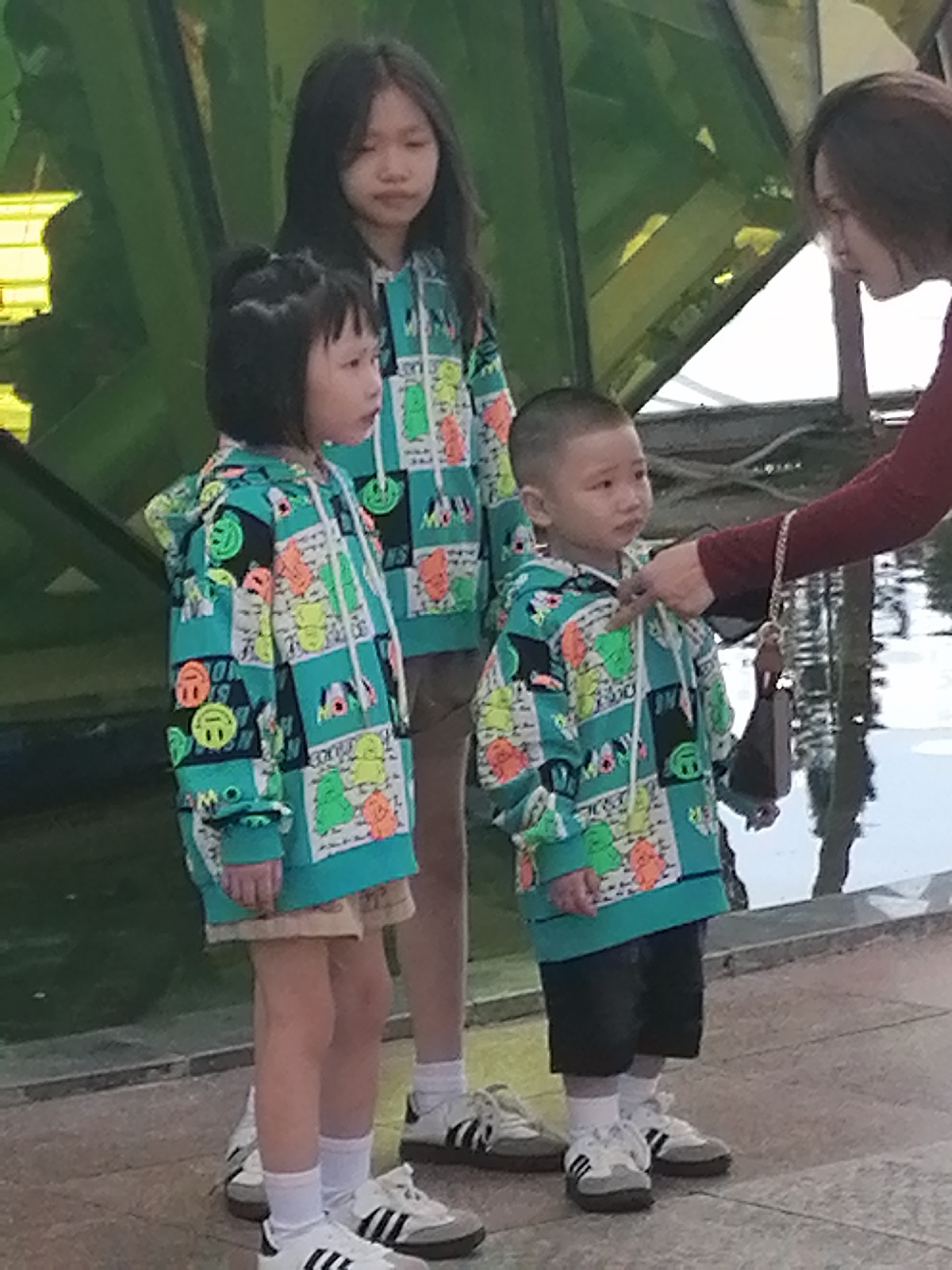 In Da Lat, Vietnam, a woman is interacting with two young children, a girl and a boy, who are standing beside an older girl. All three children are wearing matching teal hoodies with colorful cartoon prints of smiley faces and other characters. The younger girl, on the left, has her hair in a bob and is looking to her right. The boy, in the center, has a short haircut and is looking towards the woman. The older girl, behind them, has long dark hair and is looking downwards. The woman, on the right, is wearing a dark red long-sleeved shirt and is reaching out to the boy. The scene appears to be outdoors, possibly near a body of water, with a modern, angular structure in the background. The lighting suggests it is daytime.