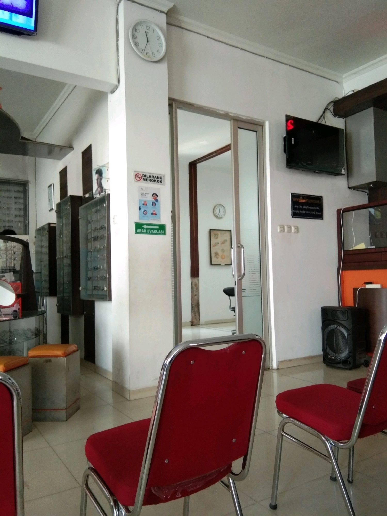The image depicts the waiting area of an optometry clinic, likely located in Kebumen, Indonesia, given the presence of Indonesian text on signage. The room is sparsely furnished with several red-upholstered chairs with chrome frames, some with plastic protection on the seats, arranged on a tiled floor. To the left, a display cabinet filled with eyeglasses is visible, suggesting the clinic's specialty. On the wall, a round analog clock indicates approximately 10:40. A flat-screen television is mounted on the wall above a darker cabinet. A sign with Indonesian text, "DILARANG MEROKOK" (No Smoking), along with a green sign indicating "ARAH EVAKUASI" (Evacuation Route) with an arrow pointing left, are prominently displayed on a white pillar. In the background, through a glass door, another room with a framed picture and a chair can be seen. A black speaker is positioned on the floor to the right of the chairs. The overall environment appears clean and functional, typical of a waiting area in a professional medical establishment.