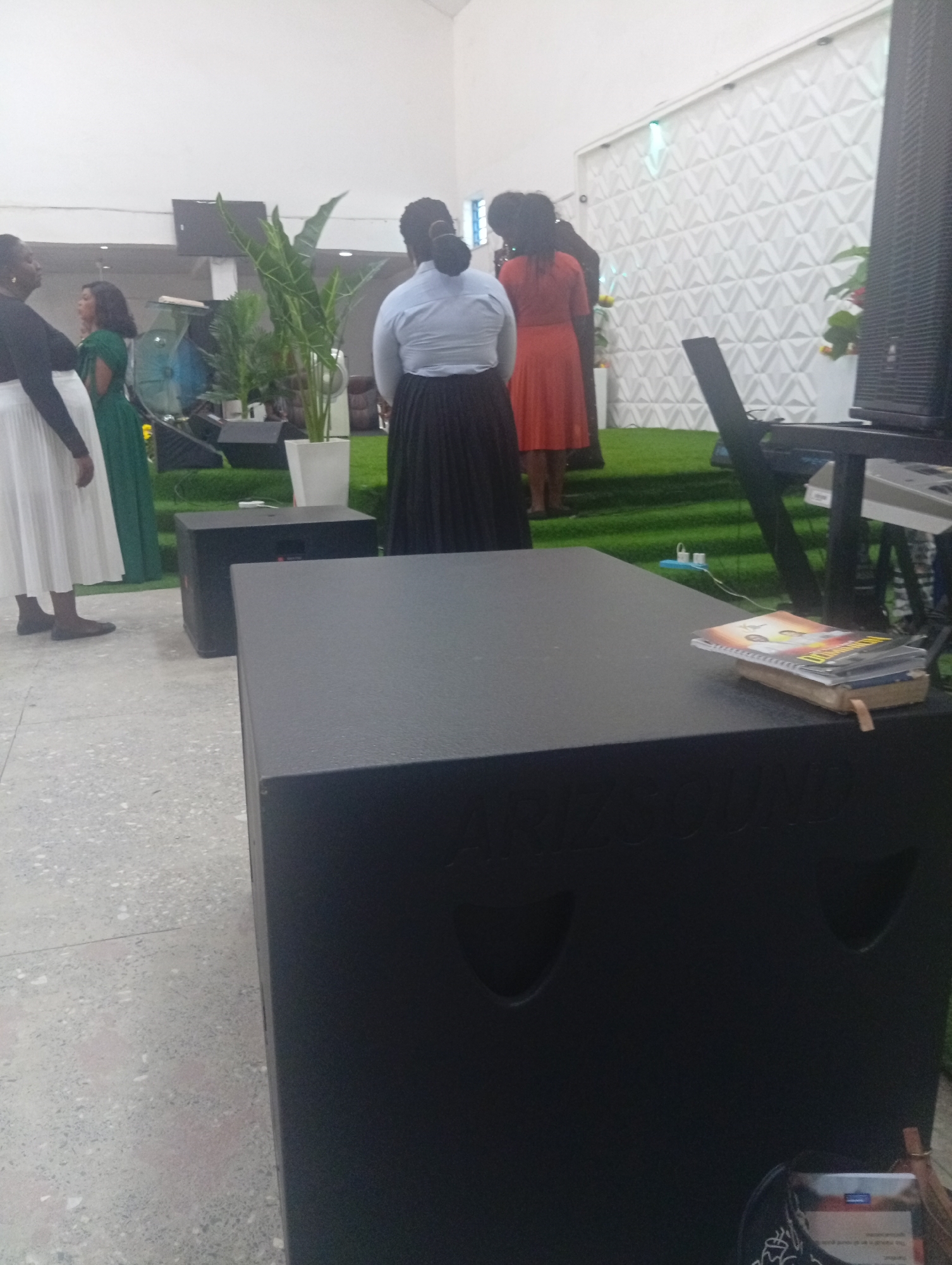 In Kason, Nigeria, inside a building with white walls and a textured geometric pattern, a group of people are gathered on artificial turf. In the foreground, a large black speaker rests on a terrazzo floor. To the left, a woman in a white skirt and black top stands next to another woman in a green dress. Behind them, several large plants in white pots are visible. Further back, a woman in a blue shirt and black skirt stands facing away from the camera, while another woman in an orange dress stands beside her. A keyboard and monitor are positioned on a stand to the right of the scene, and a black speaker is placed behind the keyboard. A stack of books or pamphlets is on top of the speaker in the foreground.