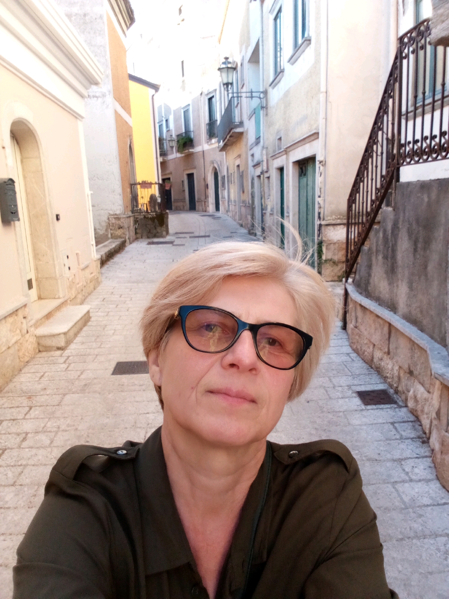 A woman with short blonde hair and black-rimmed glasses takes a selfie on a narrow cobblestone street in Castelnuovo di Conza, Italy. She is wearing a dark green collared shirt with epaulets. The street is lined with buildings, one on the left painted a pale cream with an arched entrance and a dark mailbox, and another on the right with a brown wrought-iron railing leading up a set of stairs. The buildings have a worn, aged appearance, with faded paint in shades of cream, yellow, and grey. The light suggests it is daytime, with the sun casting shadows on the cobblestones. The background shows more of the street receding into the distance, with more buildings and an old-fashioned streetlamp visible.