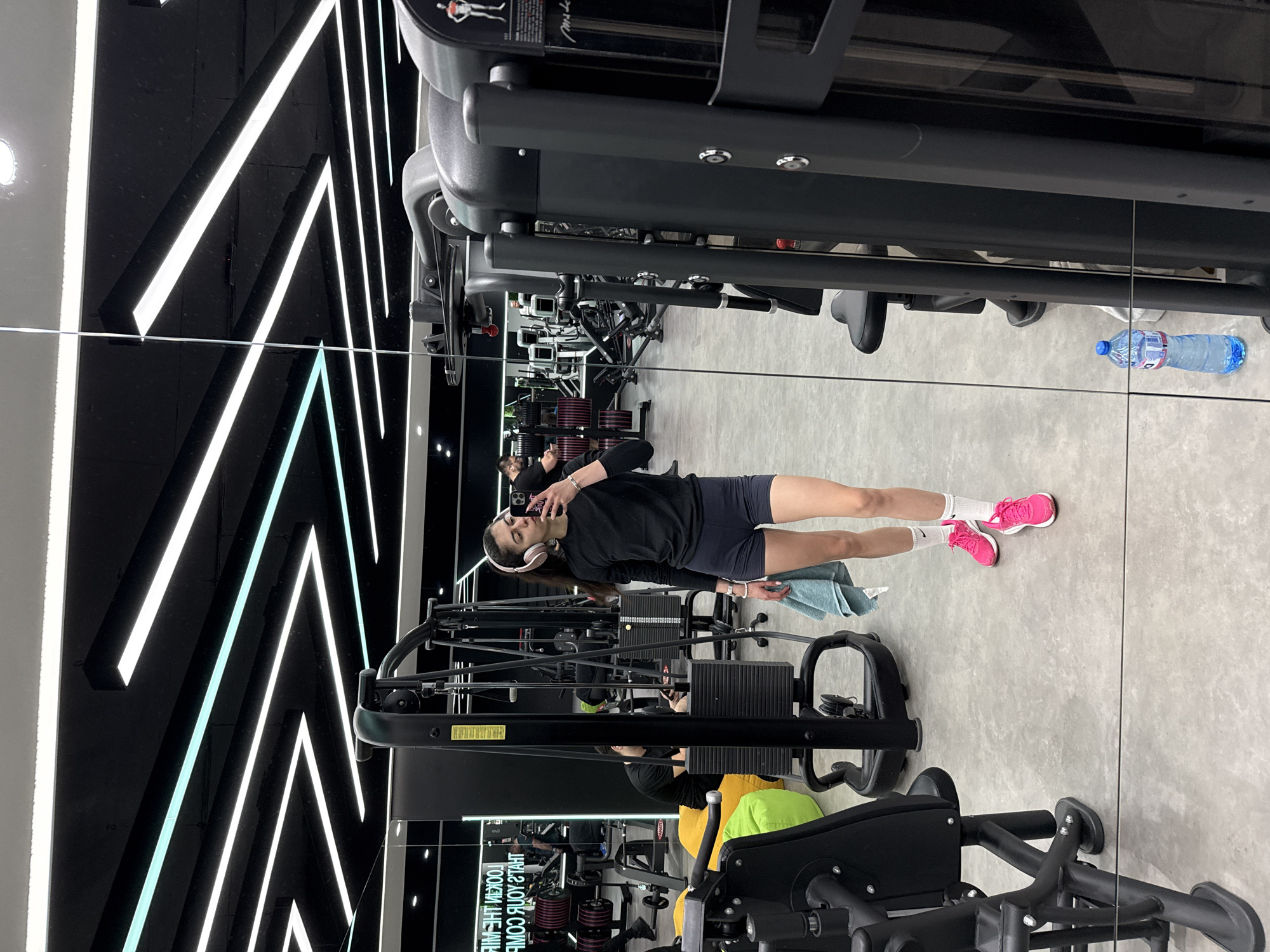 A full shot shows a woman in a gym in Sofia, Bulgaria. The woman is standing in front of a gym mirror and looking at her phone. She is wearing a black sweatshirt, dark shorts, white socks, and pink shoes. She is also wearing headphones. The gym has black walls with white neon lights and a grey floor. Gym equipment is visible around the woman, including weight machines, benches, and a water bottle. Other people are in the background.