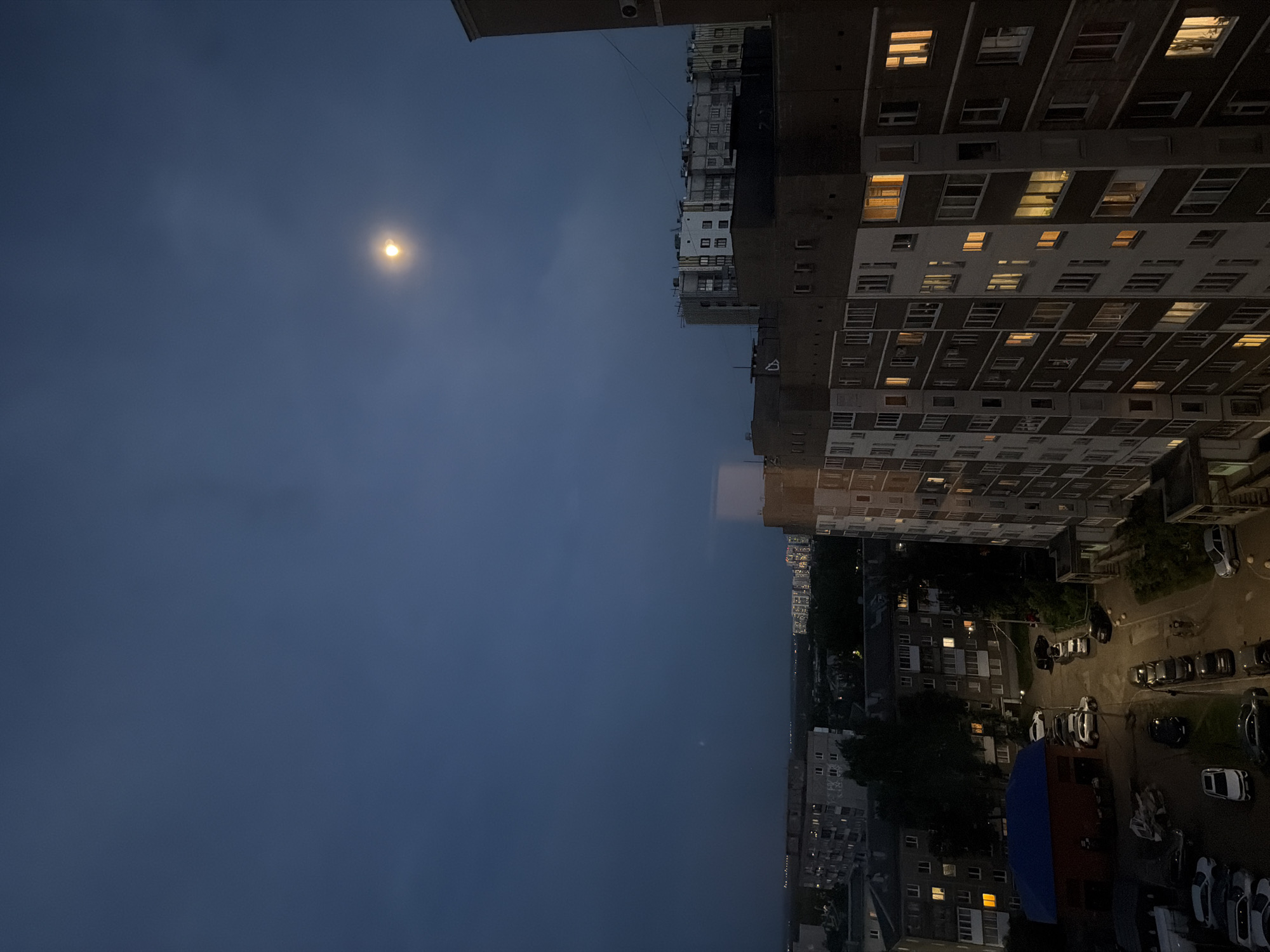 The image shows an evening view in a city, possibly in a residential area. A large building with many lighted windows dominates the right side of the frame. Below the building, a parking lot with cars is visible. Further in the distance, other buildings are illuminated, suggesting an urban landscape. The left side of the image features a dark sky with a small, bright moon. The overall mood is calm and quiet, typical of a city at night. The image has been rotated.