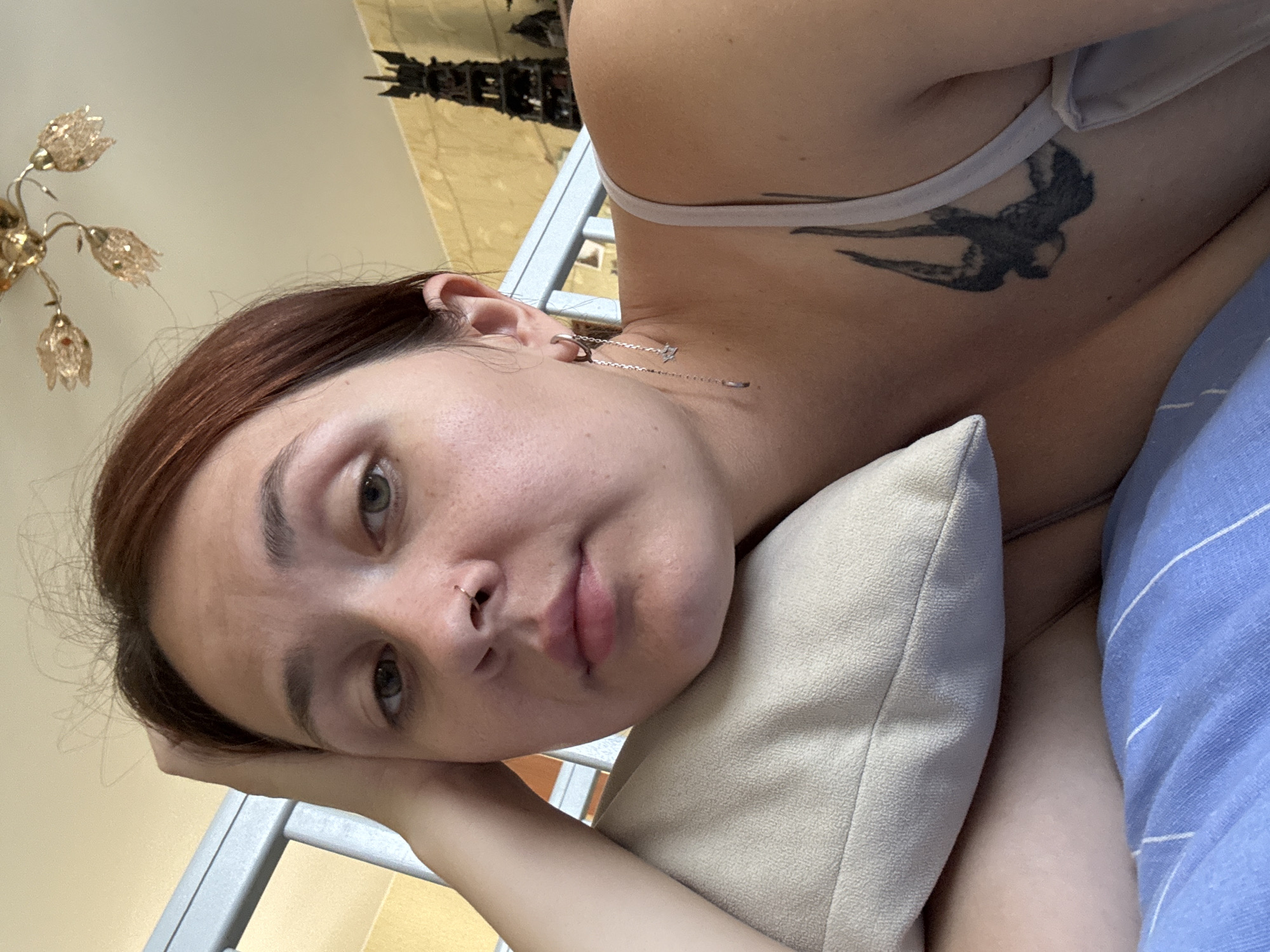 The image shows a close-up, indoor shot of a woman lying down on a bed. She has fair skin, reddish-brown hair, and is wearing a white tank top with a bird tattoo on her chest. Her left hand is resting on her head, which is supported by a light-colored pillow. The background includes a metal bed frame, a ceiling light fixture, and a decorative object on a shelf. The overall impression is one of relaxation or repose. The image does not contain any information that allows for determination of its location within the city of Naberezhnye Chelny, Russia.