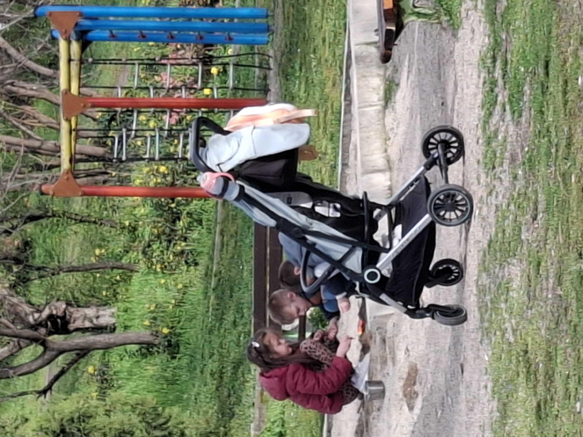 In Plovdiv, Bulgaria, a stroller stands on a paved surface near a playground and greenery. Two children, one in a red jacket and leopard print pants, and another wearing blue are on the ground near the stroller. The children are facing each other, and the girl in red appears to be interacting with something on the ground.