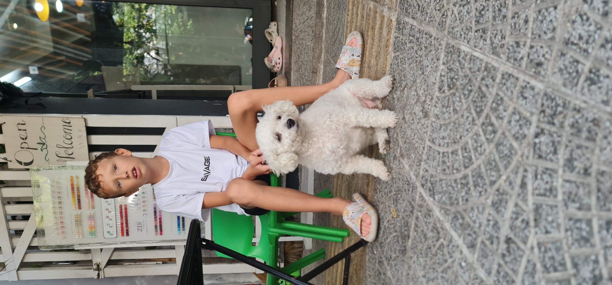A boy in a white "SAVAGE" t-shirt sits on a green chair outside a shop in Đà Nẵng, Vietnam. He holds a small, white, fluffy dog on his lap. Both are facing forward. The boy has curly, light brown hair and blue eyes. The shopfront has an "Open" sign and displays a selection of colorful items, possibly nail polish. The ground is paved with patterned grey stones. Another pair of slippers are visible on the ground behind the boy.
