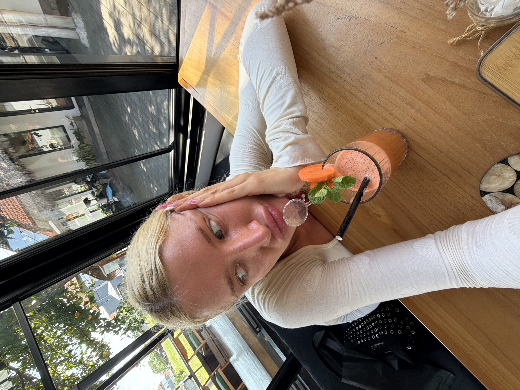 A young Caucasian woman with blonde hair sits at a wooden table in a cafe in North Kuta, Indonesia. She is wearing a white long-sleeved top. Her right hand is on her cheek, and her mouth is pouted. In front of her, on the table, is a glass of orange juice garnished with a slice of carrot and a sprig of mint, and a black straw. The background shows a view through a large window of trees, a building with a thatched roof, and other patrons in the cafe. The table also has a black bag with a pattern of silver circles on it, a small decorative item made of twine and glass, and a coaster with a mosaic pattern made of circular slices of wood.