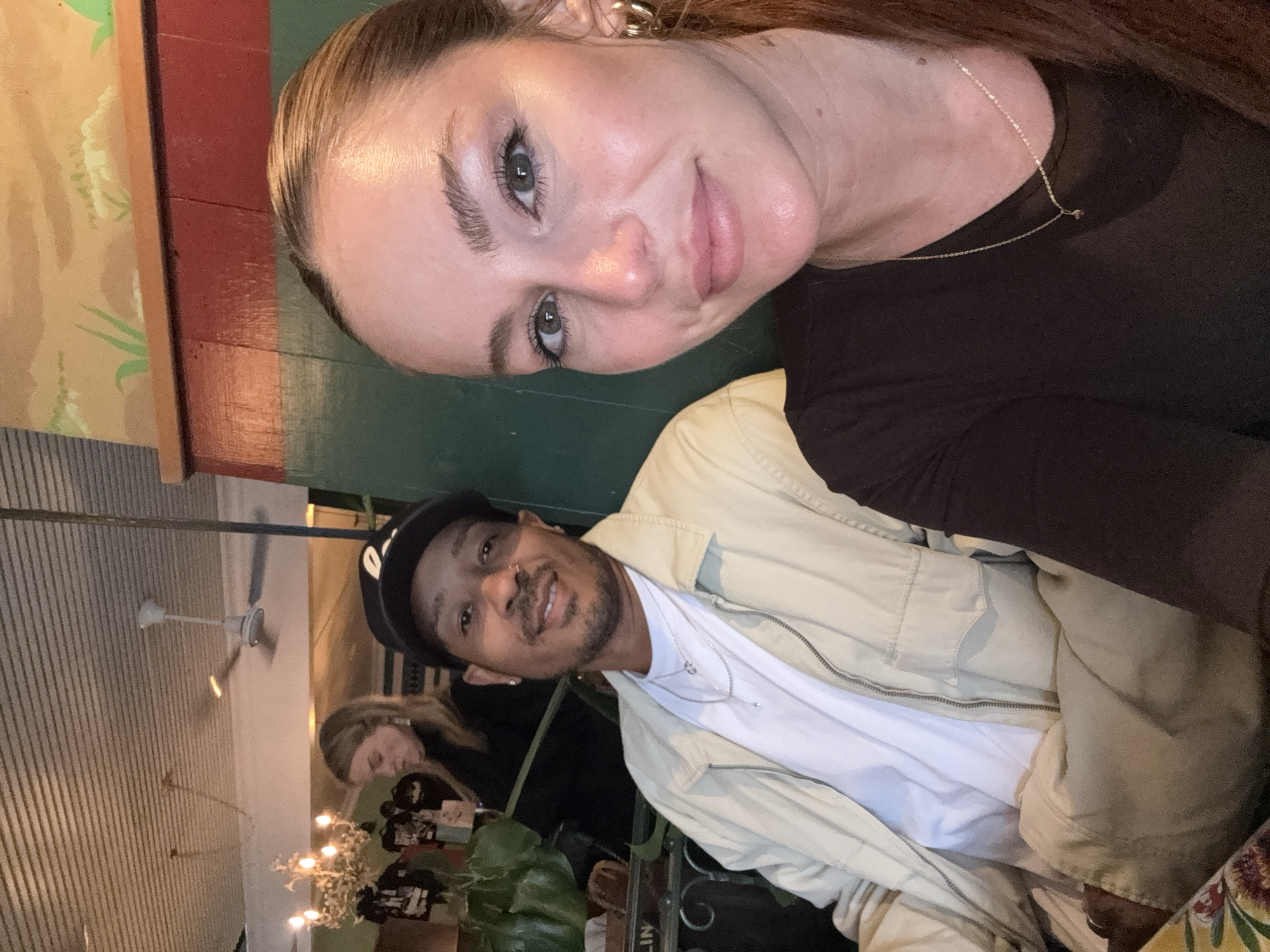 The image shows two individuals, a woman and a man, seemingly taking a selfie in what appears to be a restaurant setting. The woman, positioned on the upper part of the frame, has light skin, long brown hair, and is wearing a black top. The man, in the lower portion of the frame, has darker skin, a beard, and is wearing a black baseball cap, a white t-shirt, and a light-colored jacket.
In the background, other individuals are visible, along with interior elements of the restaurant, including plants, decorations, and a ceiling fan. The overall lighting suggests an indoor setting with warm light sources.
Without more information, it's impossible to determine the exact location in Los Angeles.