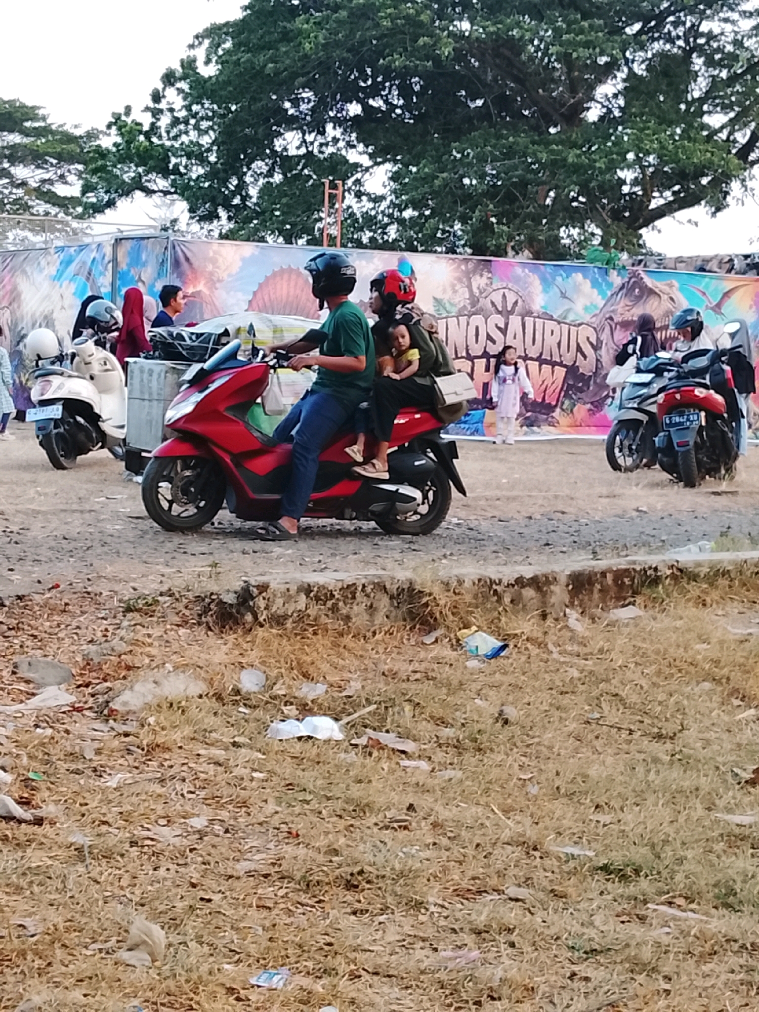 The image depicts a scene in Harjosari Lor, Indonesia, featuring several people and motorcycles in front of a large banner advertising a "DINOSAURUS SHOW." In the foreground, a person wearing a green shirt and black helmet is riding a red scooter, with a child sitting in front of them and another person, possibly a child, behind them. The rider's legs are visible, with one leg resting on the footboard. To the left, a white scooter is parked, and another person in a red headscarf is standing near it. In the background, the "DINOSAURUS SHOW" banner shows images of dinosaurs. Several other motorcycles are present, and people are gathered around. The environment appears to be an outdoor event space with a rough, dirt ground. The overall lighting suggests it is daytime.