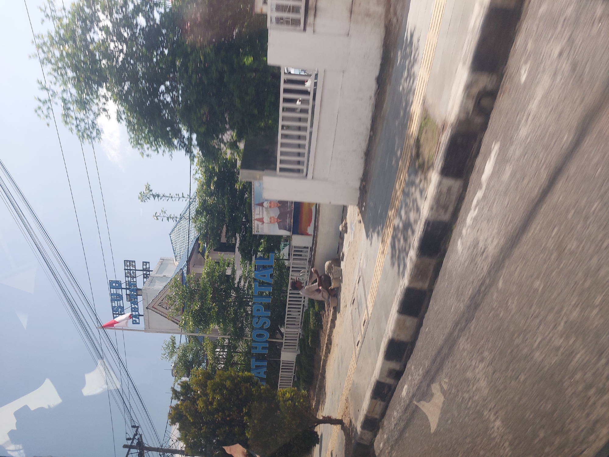 The image shows a view of a building with a sign that reads "AT HOSPITAL". A man is crouching on the sidewalk in front of the building, appearing to be working with some materials. There are trees and a fence surrounding the property. The sky is visible in the background, and there are power lines overhead. The scene appears to be in a city, possibly Parapat, Indonesia, given the context.