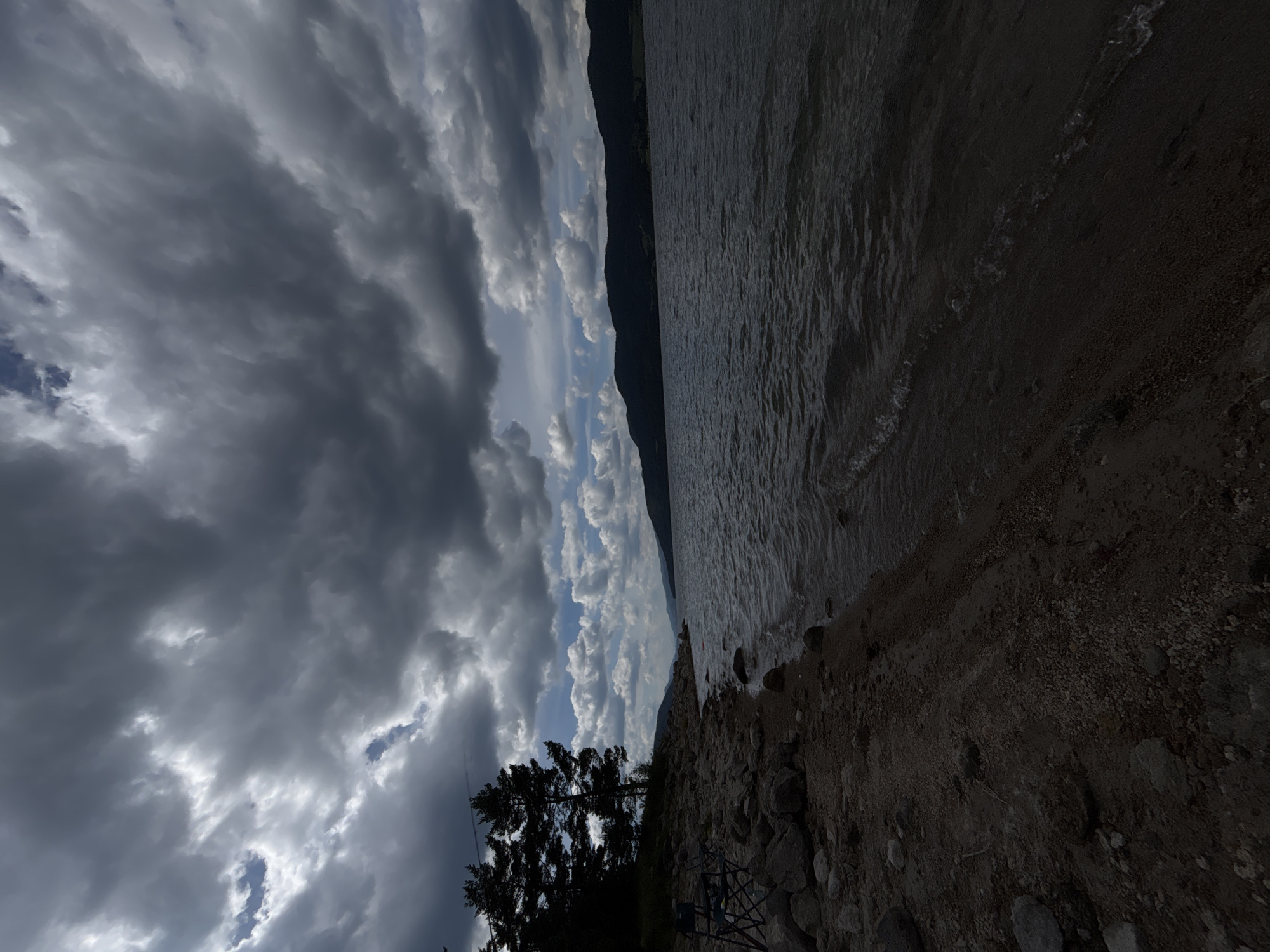 The image depicts a dark and cloudy day at a rocky lakeside. In the foreground, a sandy and pebbly shore is visible, with several rocks scattered across it. Two empty folding chairs are set up near the water's edge, suggesting a fishing or leisure activity. To the left, a cluster of dark green coniferous trees stands tall against the sky. The middle ground is dominated by a large body of dark, rippling water, reflecting the moody sky. In the distance, a silhouetted mountain range stretches across the horizon. The sky is filled with heavy, dramatic clouds, interspersed with patches of lighter, brighter clouds and hints of blue sky. The overall impression is one of a somber and perhaps slightly ominous natural landscape.