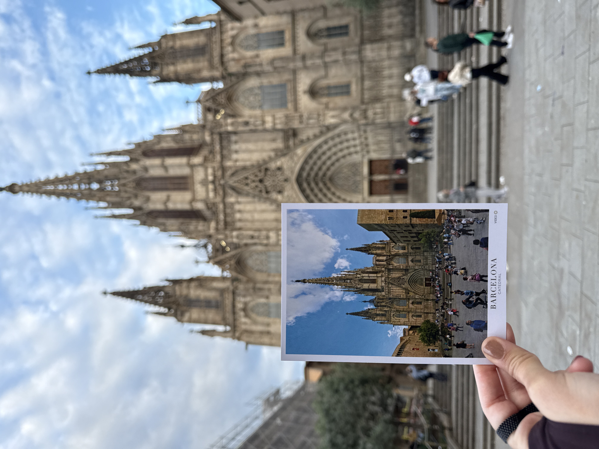 A postcard featuring a picture of the Barcelona Cathedral is held in front of the actual cathedral. The postcard image shows the cathedral with a bustling plaza full of people. The background features the cathedral’s facade in greater detail. People are walking on steps in the foreground near the edge of the frame. The sky is visible in the background. A hand holds the postcard and wears a black beaded bracelet.