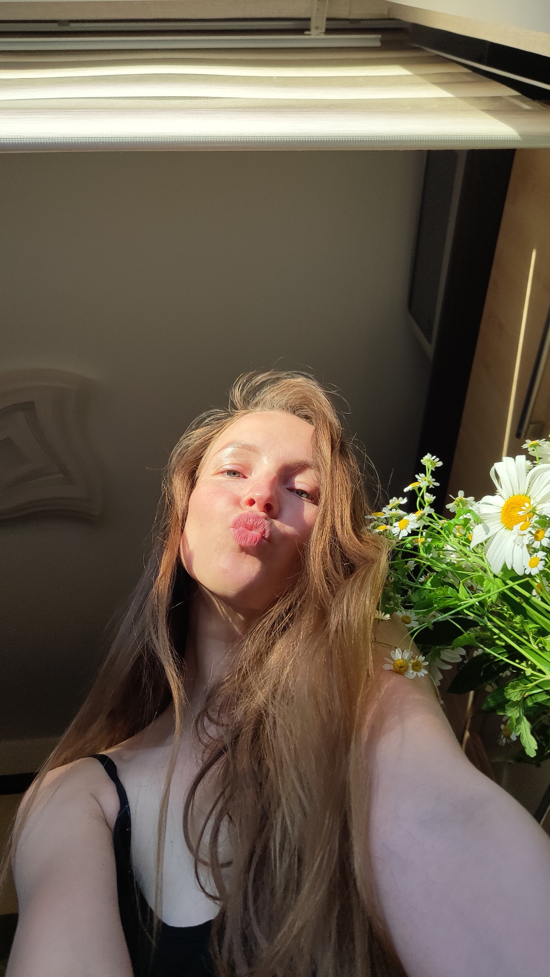 A woman with long, light brown hair puckers her lips in a selfie. She is wearing a black tank top. Beside her is a bouquet of white daisies. The background includes a window with a closed white blind, and a cream-colored wall. The image is taken indoors with sunlight casting shadows on the subjects. There is no information about the city of Chaikovsky, Russia.
