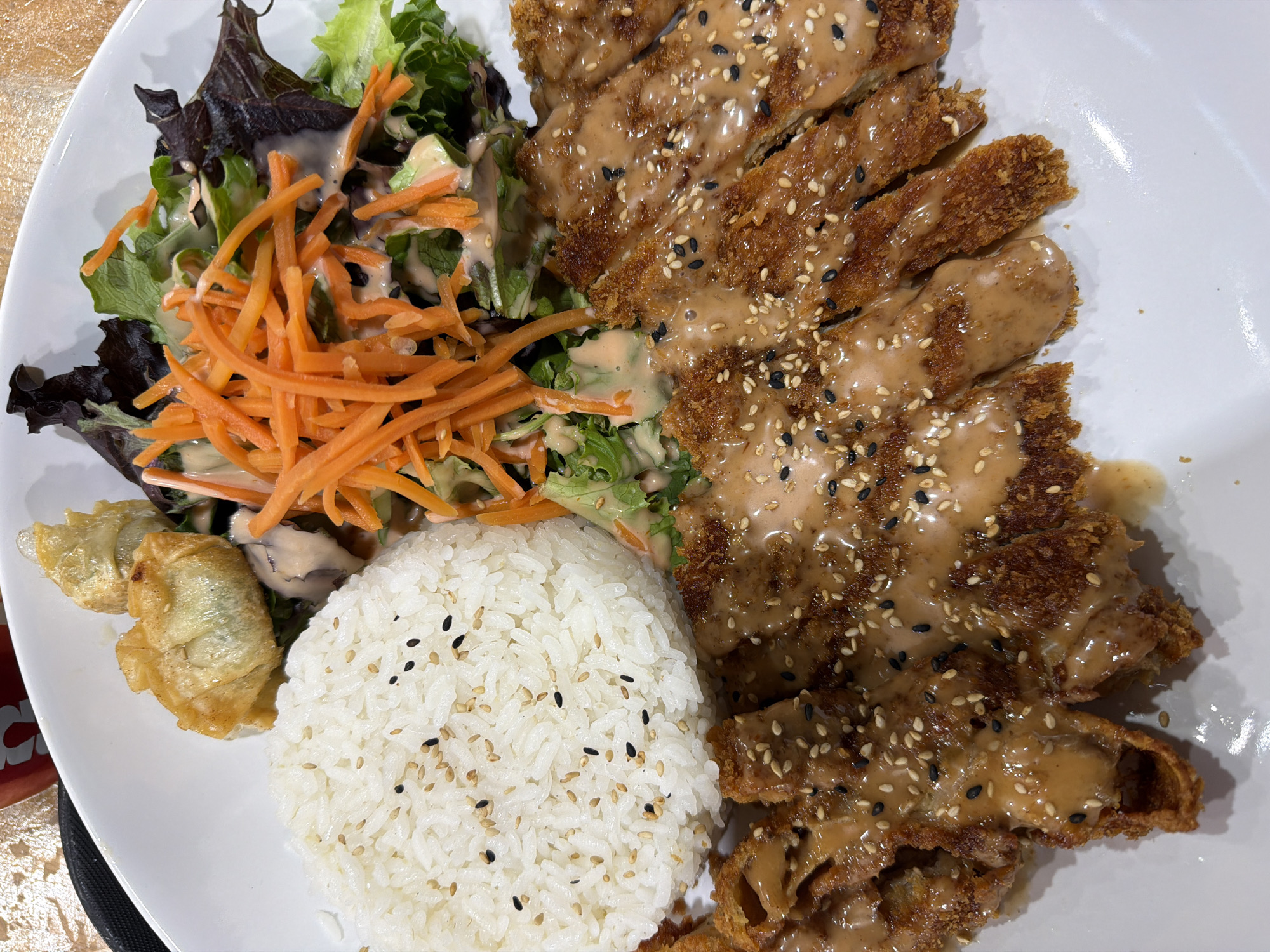 On a white plate, there is a presentation of a meal consisting of sliced, breaded, and fried meat, rice, salad, and dumplings. The meat is coated with a light brown sauce and sprinkled with sesame seeds. The rice is white and also speckled with sesame seeds. The salad includes green lettuce, shredded carrots, and a light-colored dressing. Next to the rice are two fried dumplings. The location is Niles, United States of America.