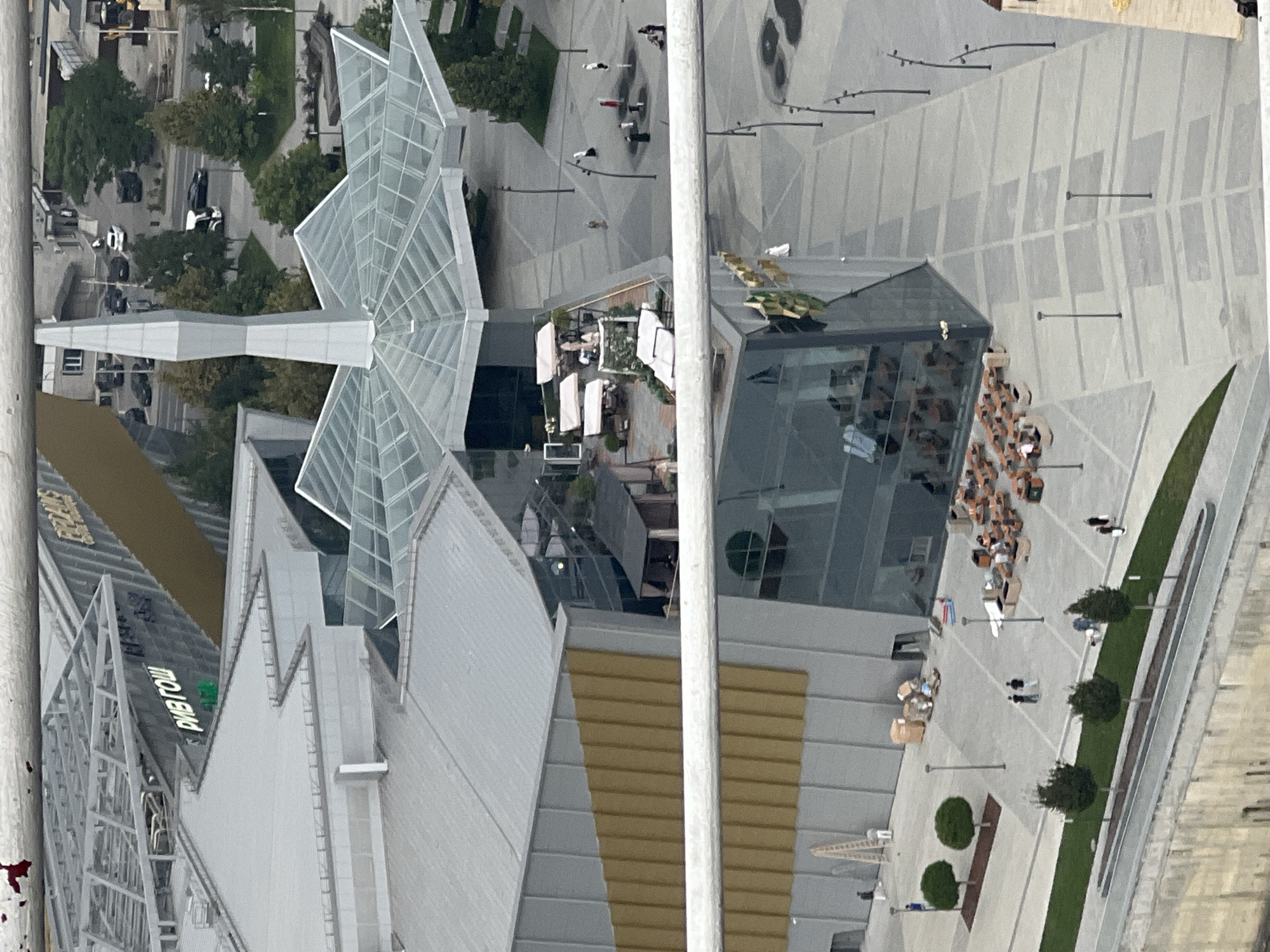 This is an aerial, high-angle, medium shot of an outdoor plaza in Grozny, Russia, featuring modern architecture and public spaces.
In the foreground, a section of a building with a glass facade and a golden-colored exterior panel is visible. Outside this building, several outdoor seating areas with tables and chairs are set up, some covered by umbrellas. A few people are seated at these tables.
Moving further into the scene, there is a large, open plaza with a patterned pavement. Several lampposts are spaced throughout the plaza. A few individuals are walking or standing in the plaza. There are also some small trees and green spaces.
In the background, a larger, complex structure with a distinctive, multi-faceted glass roof and a tall, pointed spire is prominent. Part of a building with the Cyrillic text "РИВ ГОШ" is also visible, indicating a store or shopping center. The surrounding area appears to be urban, with roads and trees visible in the distance, along with parked vehicles.
The overall scene suggests a contemporary urban environment with public amenities designed for gathering and leisure.