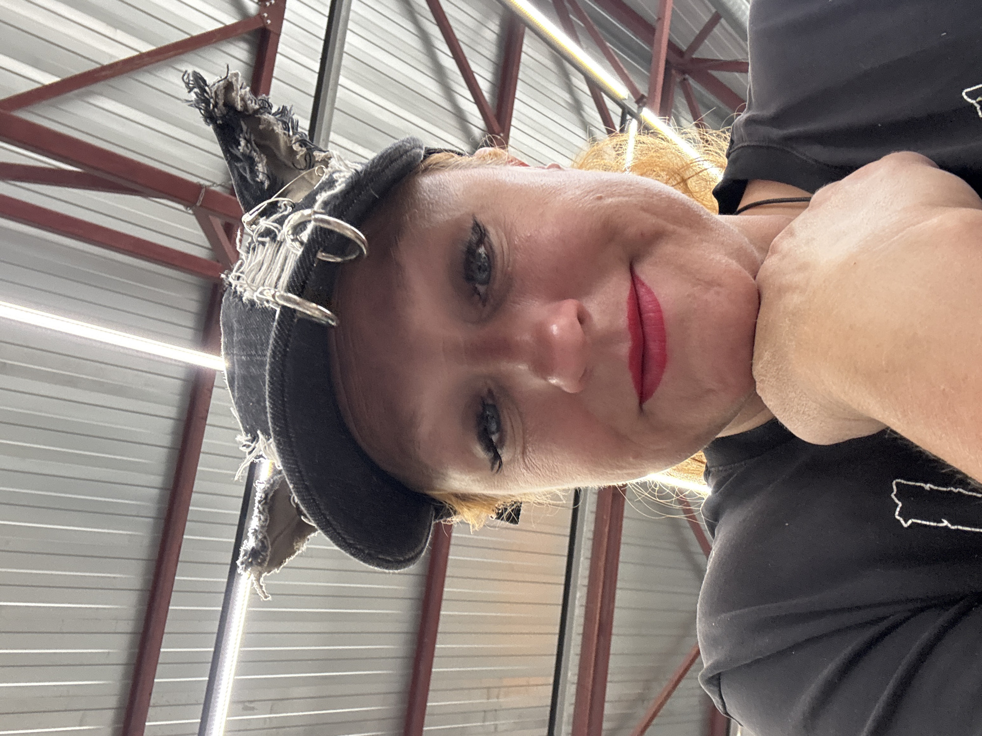 A low-angle, close-up selfie shows a woman looking down towards the camera with a calm, slightly smiling expression. Her fair skin, light-colored (possibly red or auburn) hair, dark eye makeup, and bright red lipstick are prominent. She is wearing a dark, distressed fabric hat adorned with multiple silver rings and safety pins, and a black t-shirt. On her left shoulder, a white outline graphic resembling a landmass or state is visible on the t-shirt.
The image is captured indoors, with the background revealing the ceiling and upper walls of what appears to be an industrial or warehouse-like building in Chaikovsky, Russia. The ceiling features corrugated metal panels, supported by prominent red structural beams. A linear light fixture is also visible in the upper right portion of the frame, providing bright illumination. The overall setting suggests a practical, possibly working environment. No specific weather conditions can be discerned from this indoor setting. There is no discernible text beyond the graphic on the t-shirt.