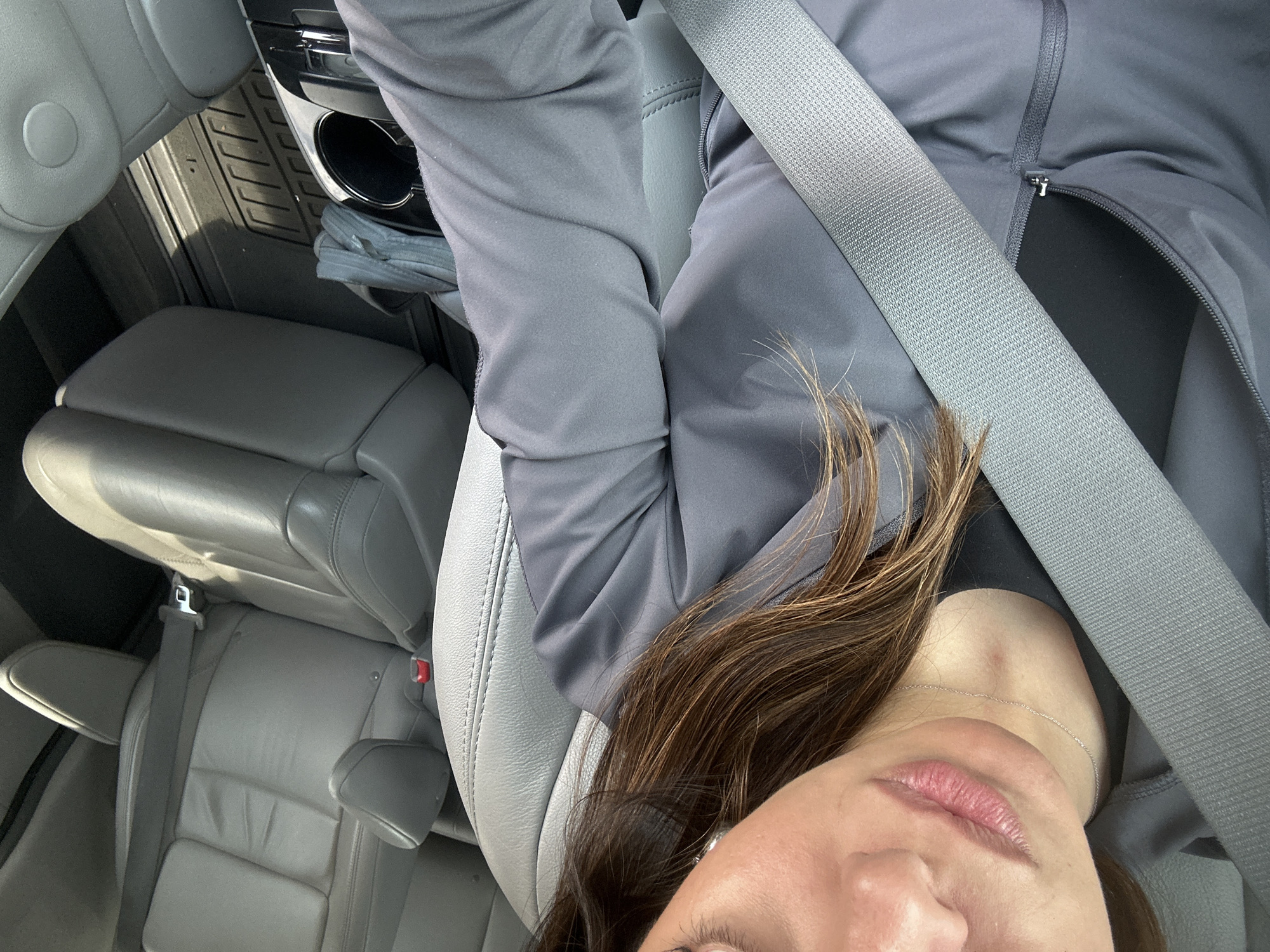 The image is a rotated, upward-angled view from inside a car, likely a selfie taken by the driver. The primary subject is a person, partially visible, with brown hair and fair skin, wearing a gray zip-up jacket and a thin silver chain necklace. Their lips are slightly parted, and part of their eye is visible at the bottom of the frame. A gray seatbelt is fastened across their chest and shoulder.
The setting is the interior of a car, characterized by light gray leather seats. The driver's seat, where the person is sitting, has visible stitching. In the background, beyond the person's arm, black, textured rubber floor mats are visible, stretching across the footwell area. A cup holder console is partially seen to the person's right. Further back, two rows of passenger seats, also in light gray leather, are discernible, suggesting a larger vehicle like an SUV or minivan.
Strong, natural light enters the car, casting shadows, indicating it is daytime, likely sunny. No specific external location cues are visible. There is no visible text in the image.